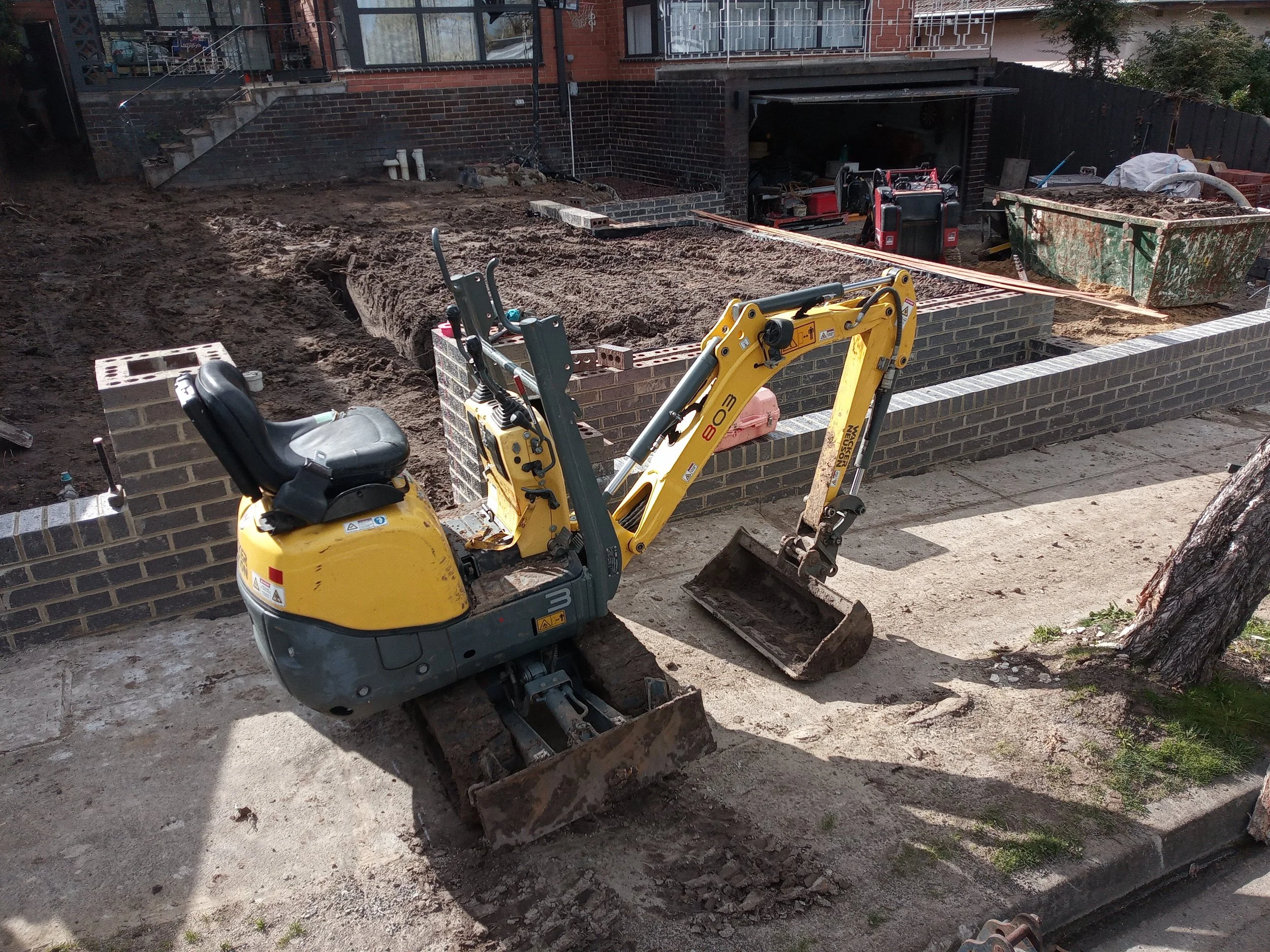 Small yellow excavator on a dirt construction site, with partially built brick wall and construction materials in background.