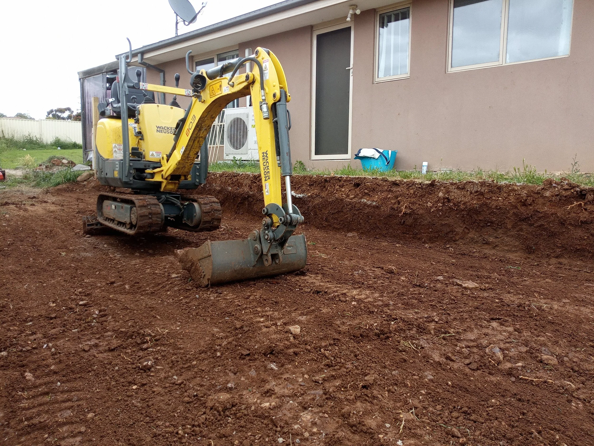 Small yellow excavator on a dirt lot in front of a house, with a blue container and a white bottle nearby.