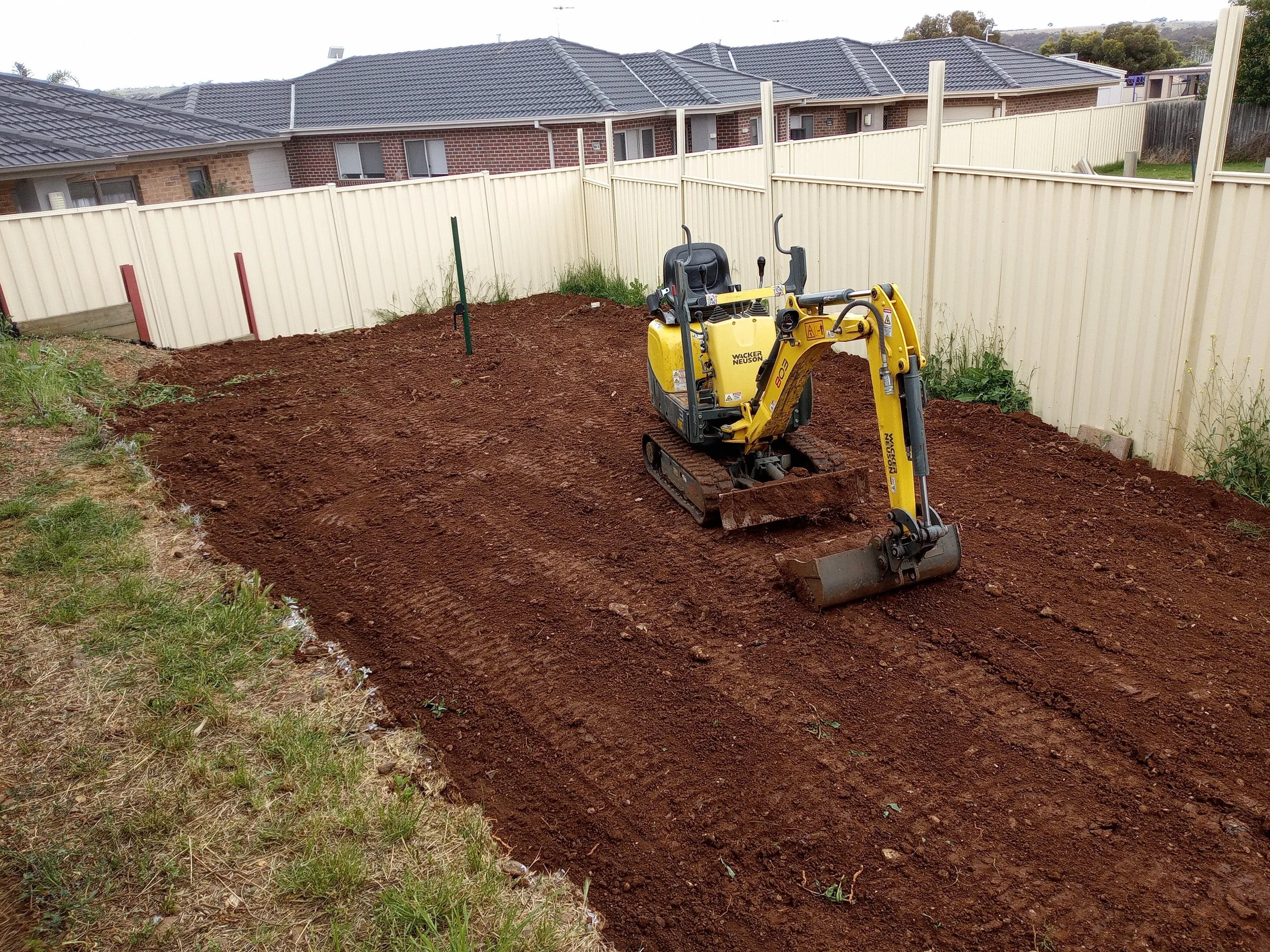 A small yellow excavator working on a backyard construction site, leveling the soil, surrounded by a white picket fence with neighboring houses in the background.
