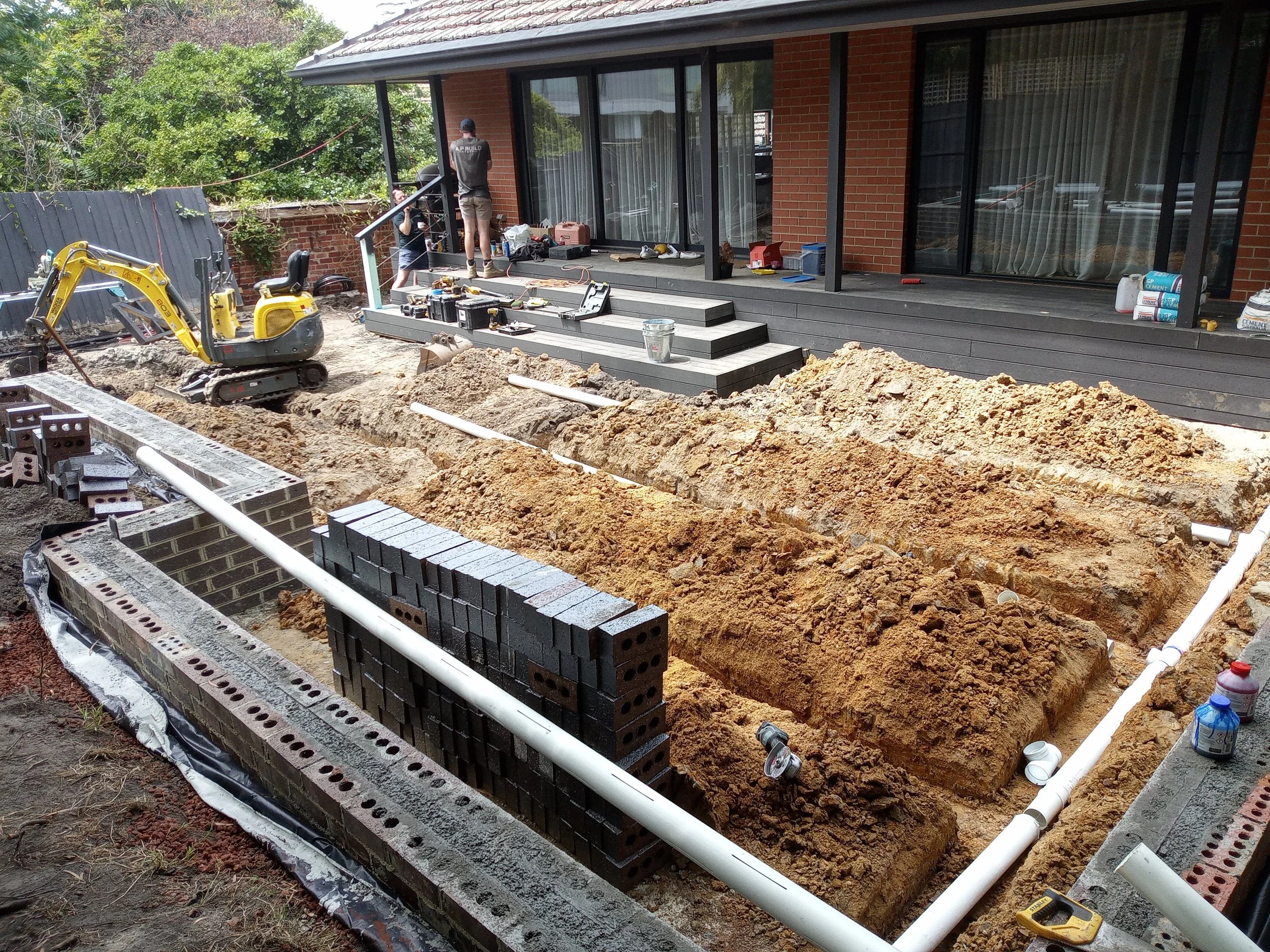 Construction site in backyard with trench for plumbing, a small yellow excavator, building materials, and workers installing infrastructure.