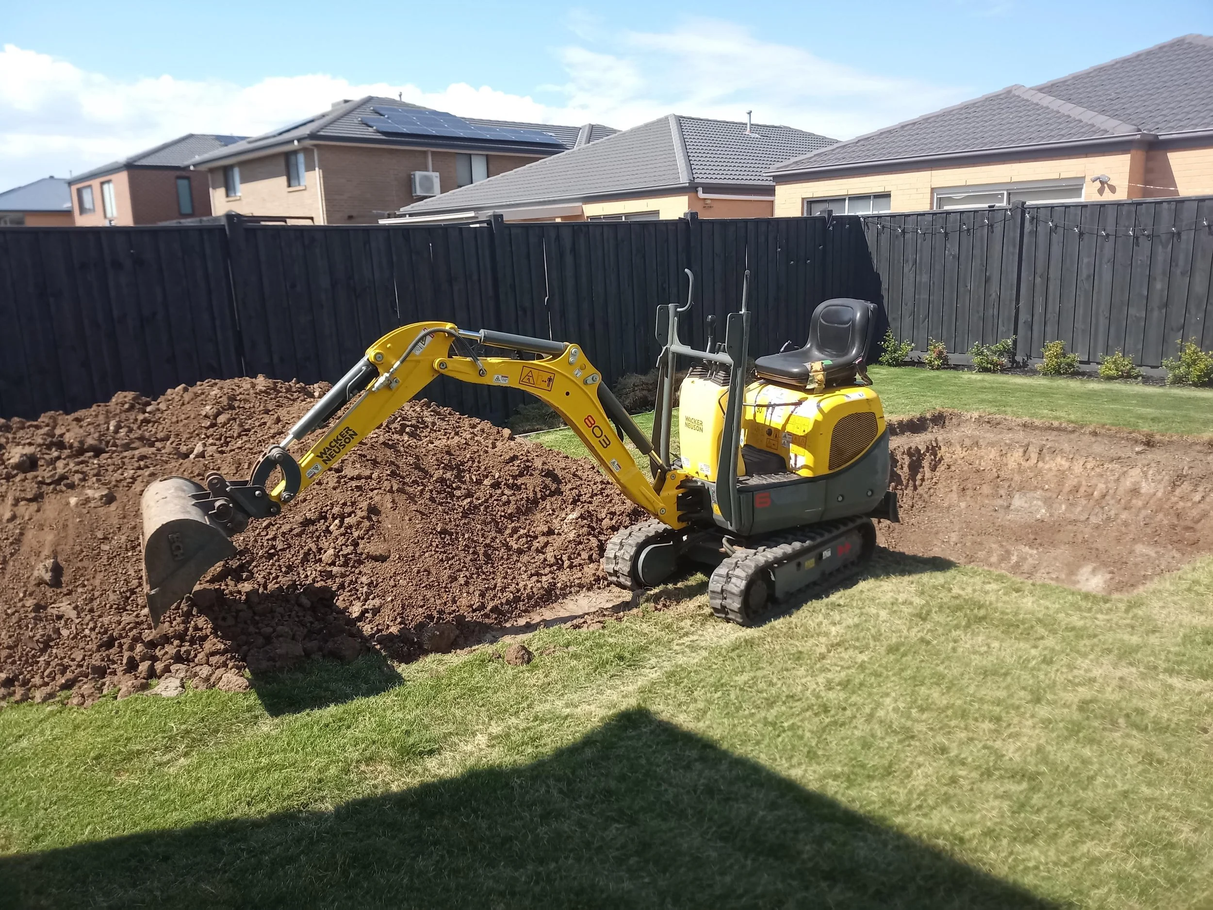 Mini excavator digging a hole in a backyard with houses and a black fence in the background.