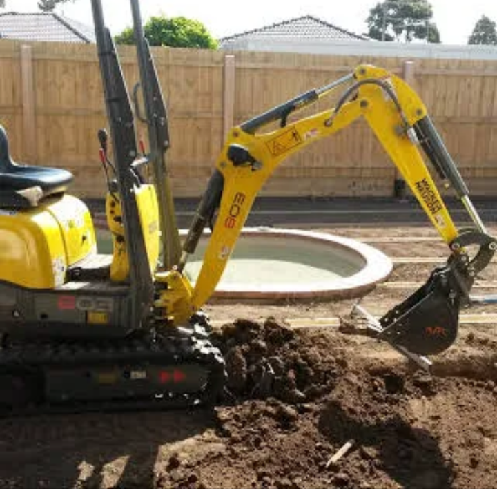 Small yellow excavator digging in a backyard with a circular plastic pool and a wooden fence.