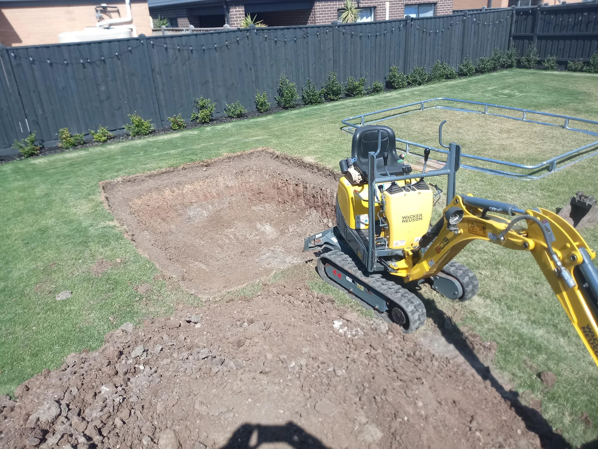 A small yellow excavator digging a square hole in a backyard with green grass, a black fence, and some plants along the fence line.