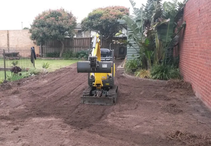 A small yellow compact excavator is digging and leveling soil in a backyard, with a brick wall on the right and a fence with trees in the background.
