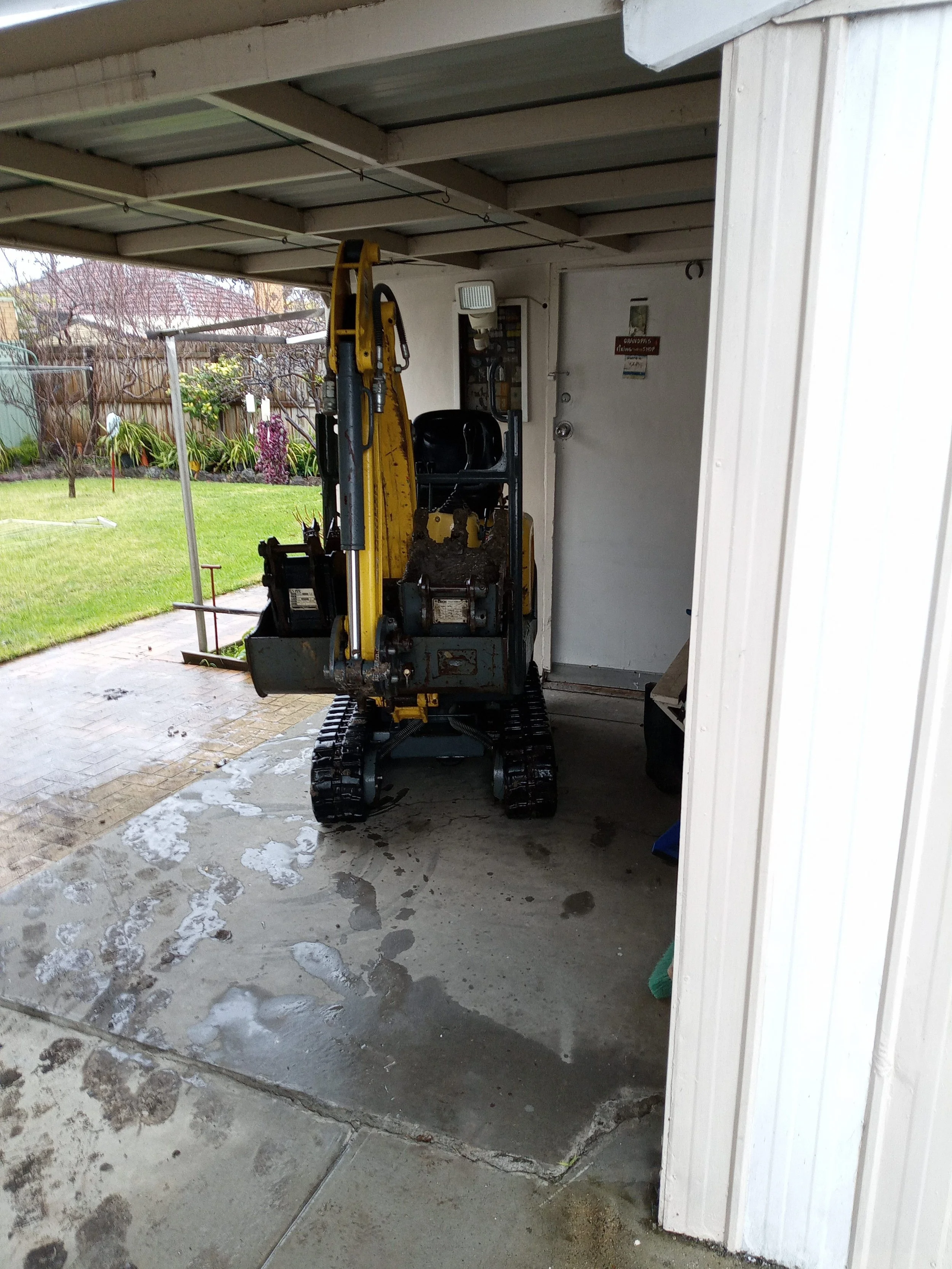 A small yellow and black mini excavator parked inside a garage next to a white door, with wet concrete on the ground and a view of a backyard with grass and trees outside.
