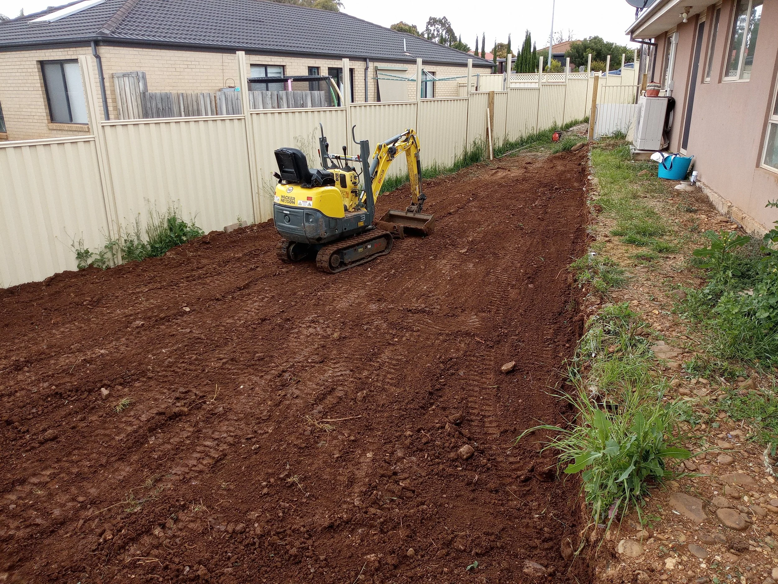 A small yellow excavator on a dirt patch in a backyard, with a white fence, neighboring houses, and some plants along the edge.
