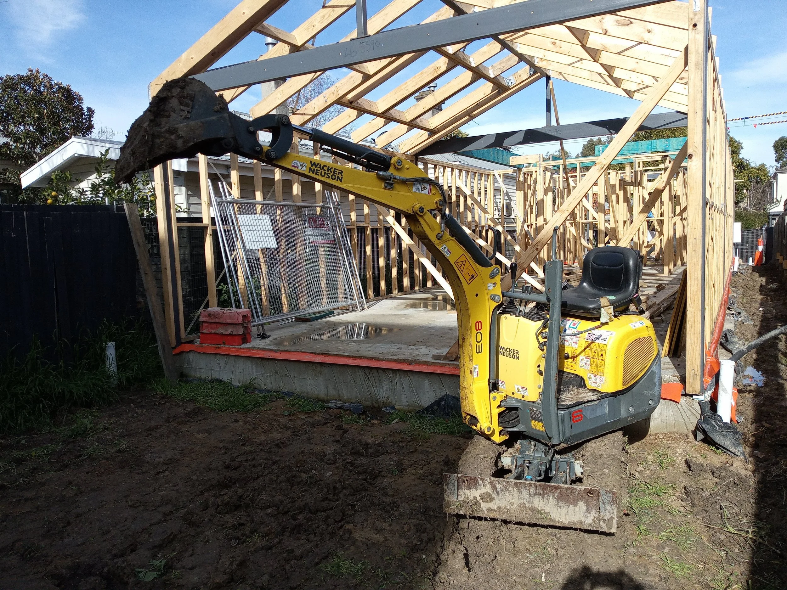 A construction site with a small yellow excavator in the foreground and a partially built wooden structure in the background.