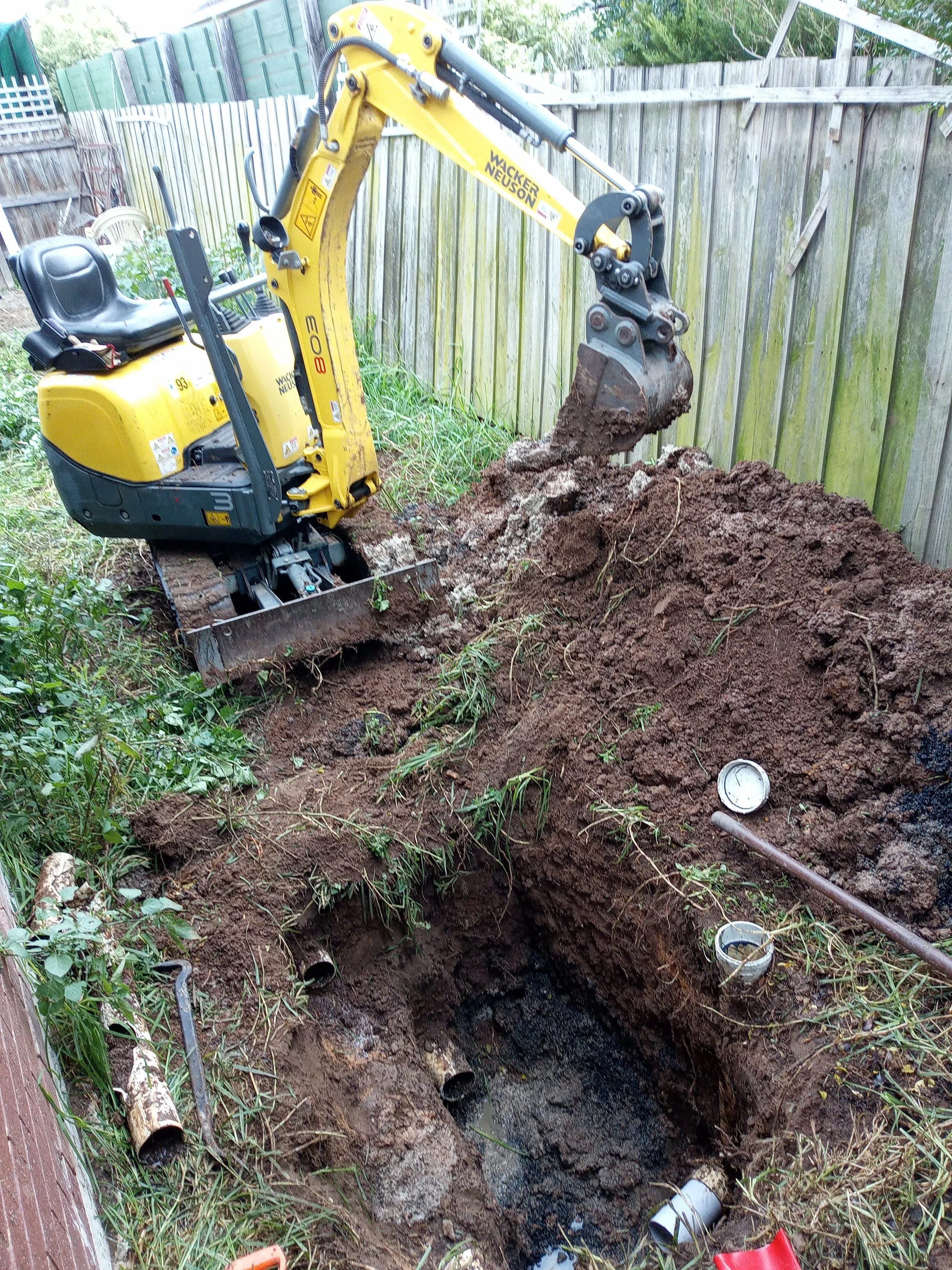 A small yellow excavator digging a trench in a backyard with a wooden fence in the background. The trench contains pipes and some water at the bottom.