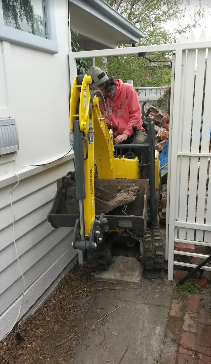 A person operating a small yellow excavator with a bucket filled with dirt or debris in a confined outdoor space next to a house with white siding, a white fence, and some brickwork.
