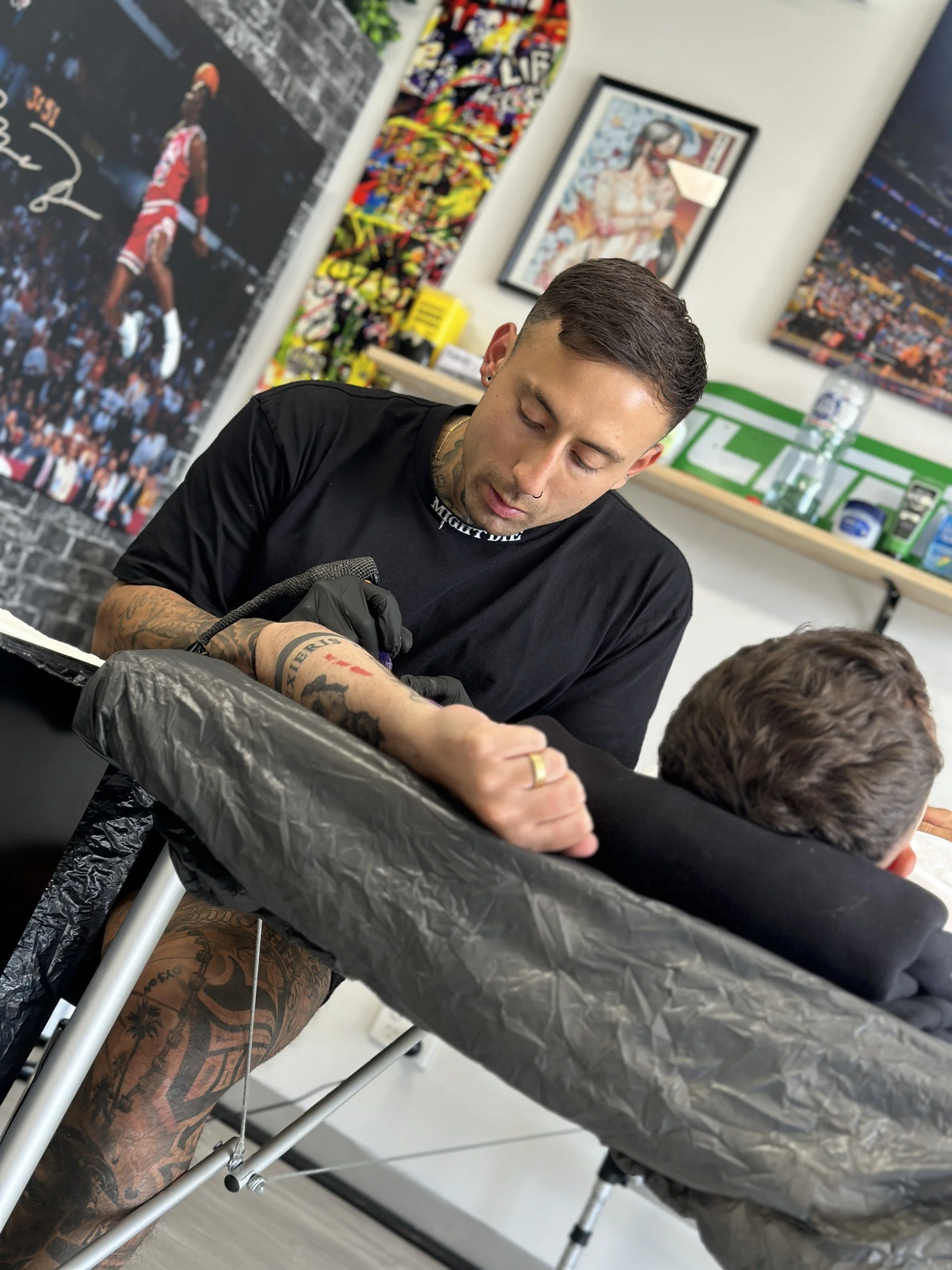 Tattoo artist working on a client in a tattoo parlor, surrounded by various artwork and posters on the walls.
