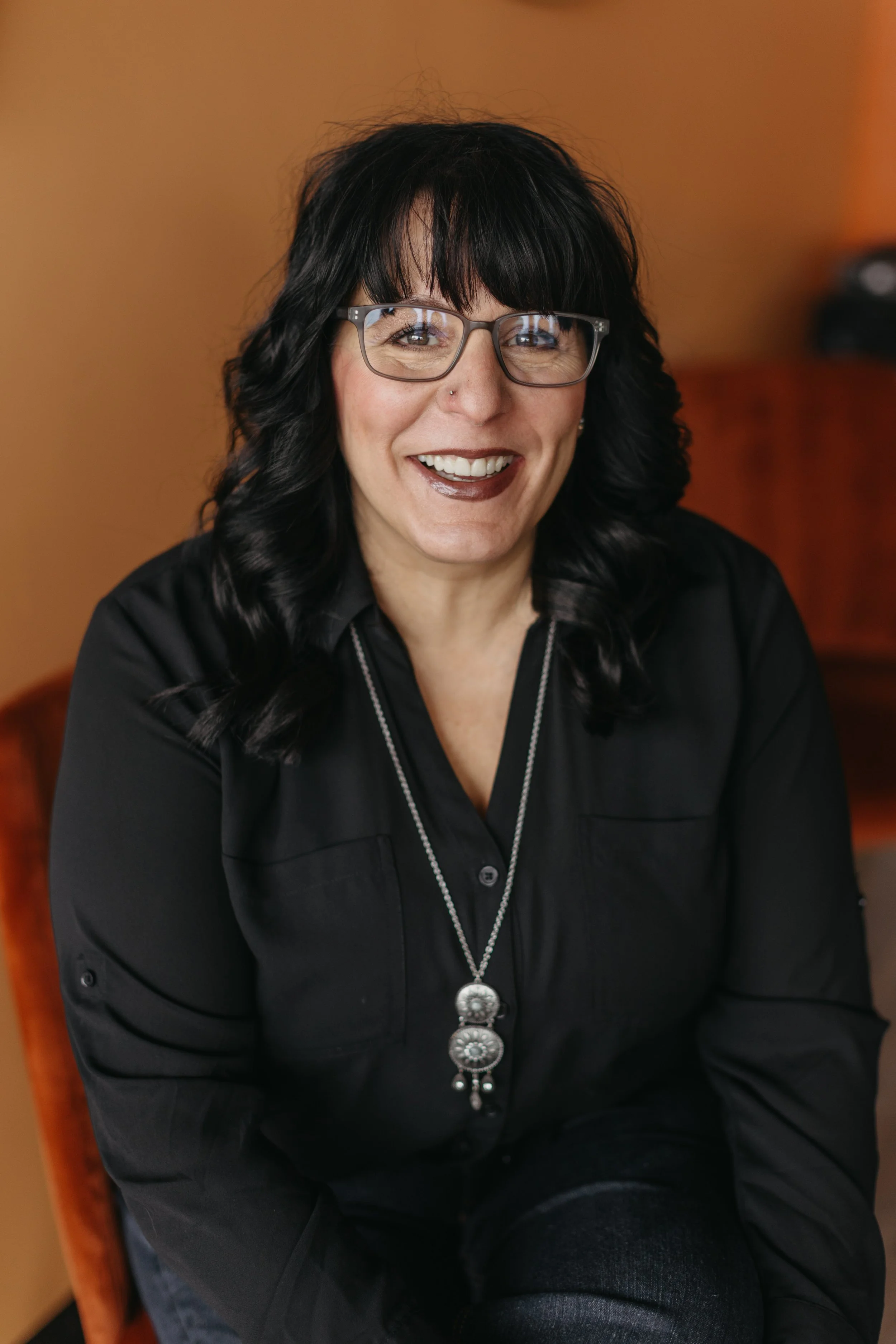 A woman with black hair, glasses, and dark lipstick smiles while sitting in a room with warm-colored walls.