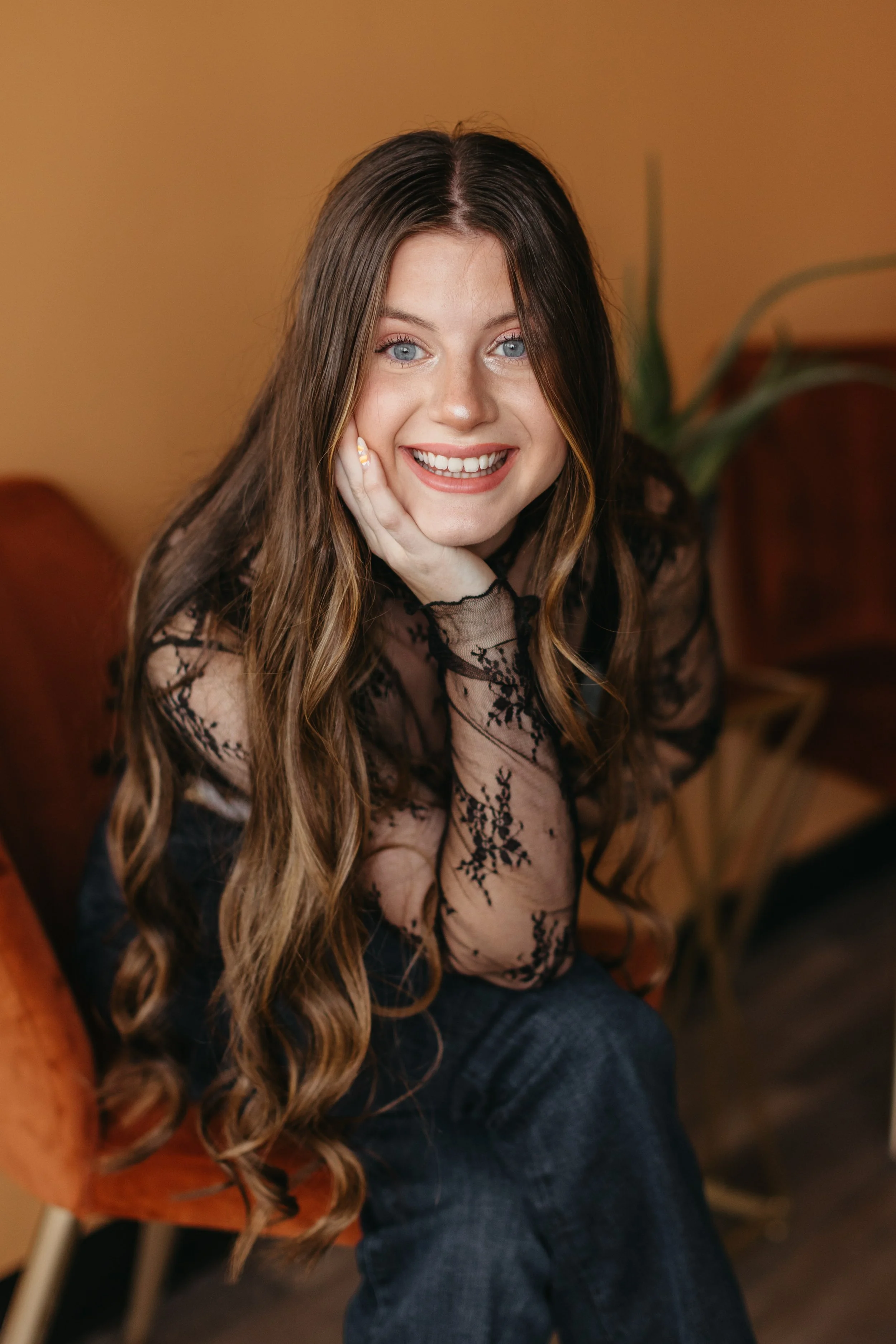 A young woman with long wavy brown hair and blue eyes sitting on an orange chair, smiling with her hand resting on her chin, wearing a sheer black lace top with floral patterns and blue jeans.