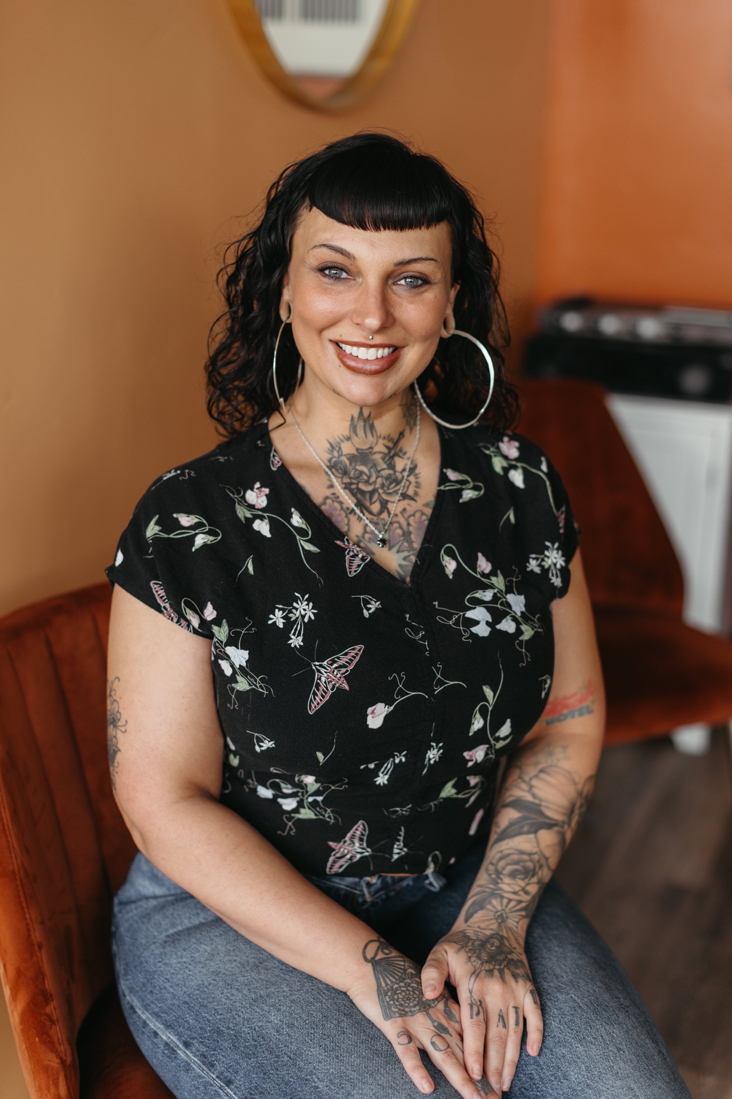 A woman with black curly hair and tattoos on her arms and chest, sitting on an orange velvet chair, smiling at the camera. She is wearing large hoop earrings and a black floral top.
