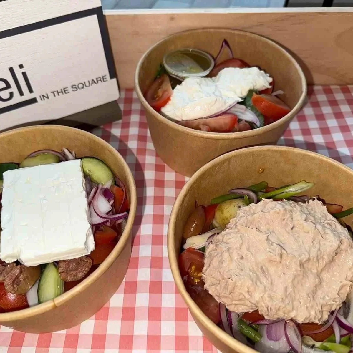 Three bowls of salad with vegetables, feta cheese, and toppings such as chicken or tuna, on a checkered tablecloth.