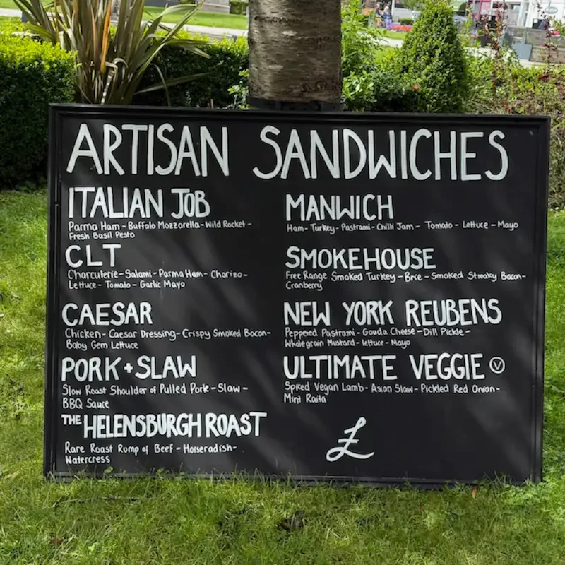 Outdoor menu board listing various artisan sandwiches including Italian Job, CLT, Caesar, Pork & Slaw, The Helensburgh Roast, Manwich, Smokehouse, New York Reubens and Ultimate Veggie, with ingredients and descriptions.