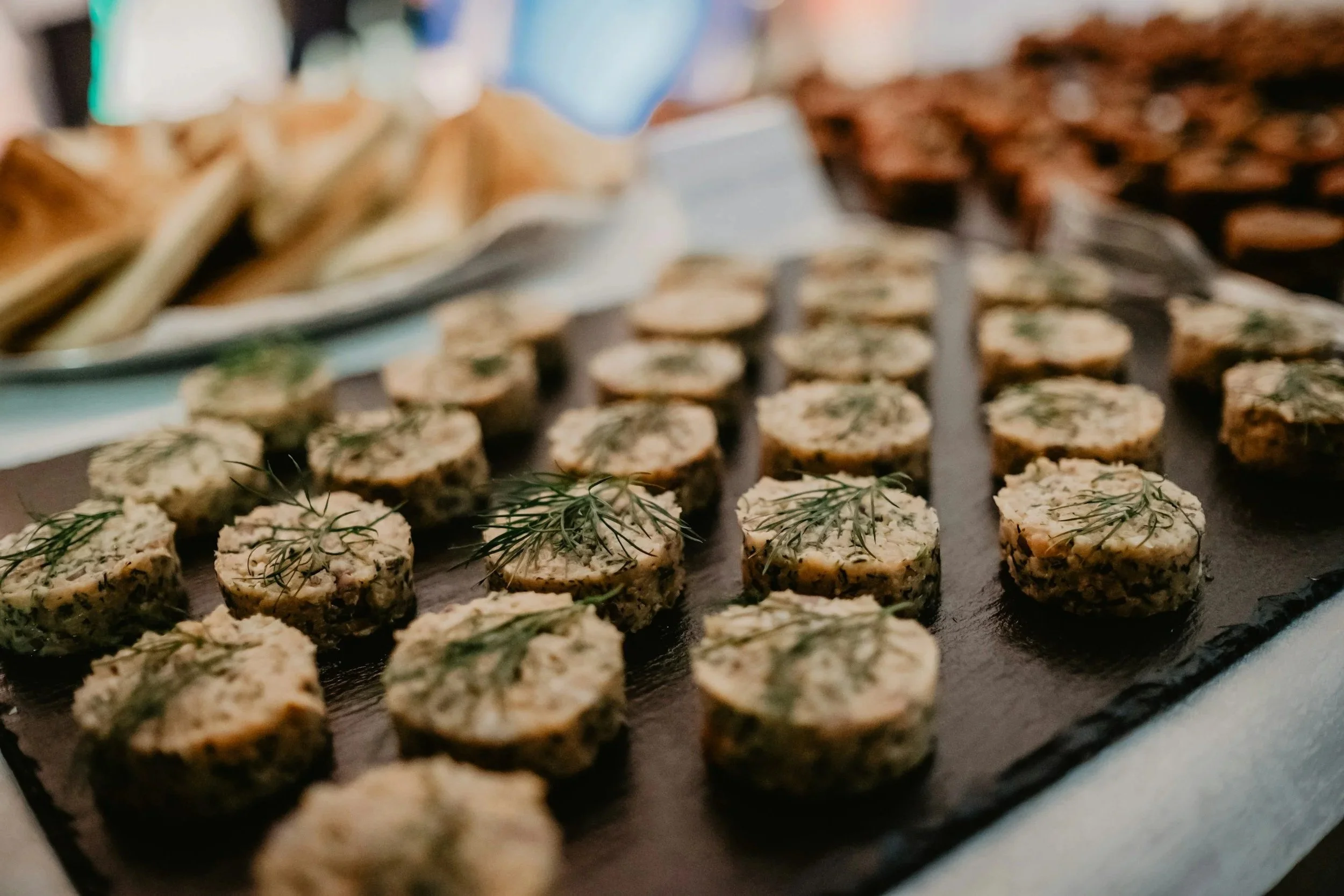 A slate platter of circular canapés with plates of food blurred in the background.