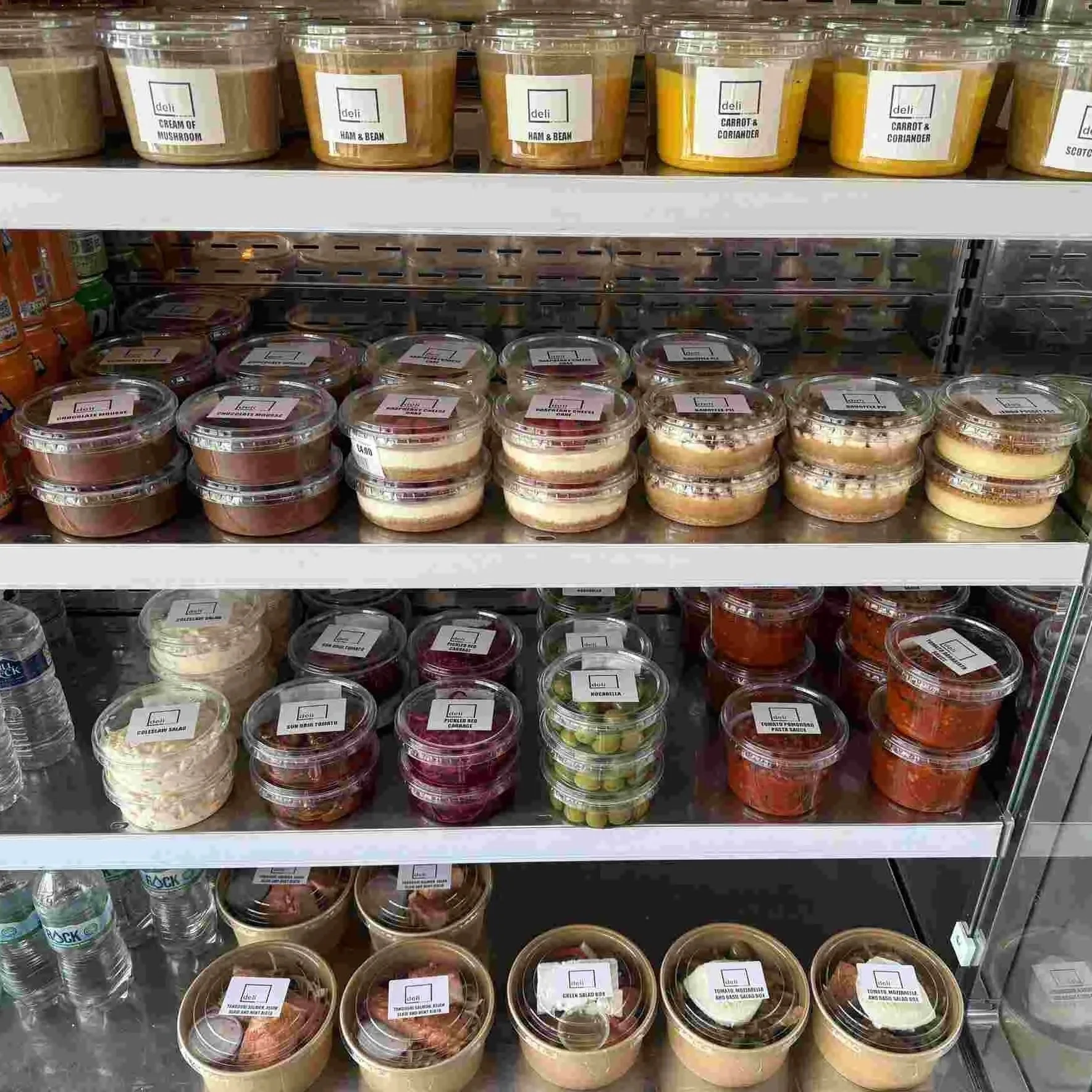 Shelf of pre-packaged salads, desserts, and some bottled water in a grocery store.