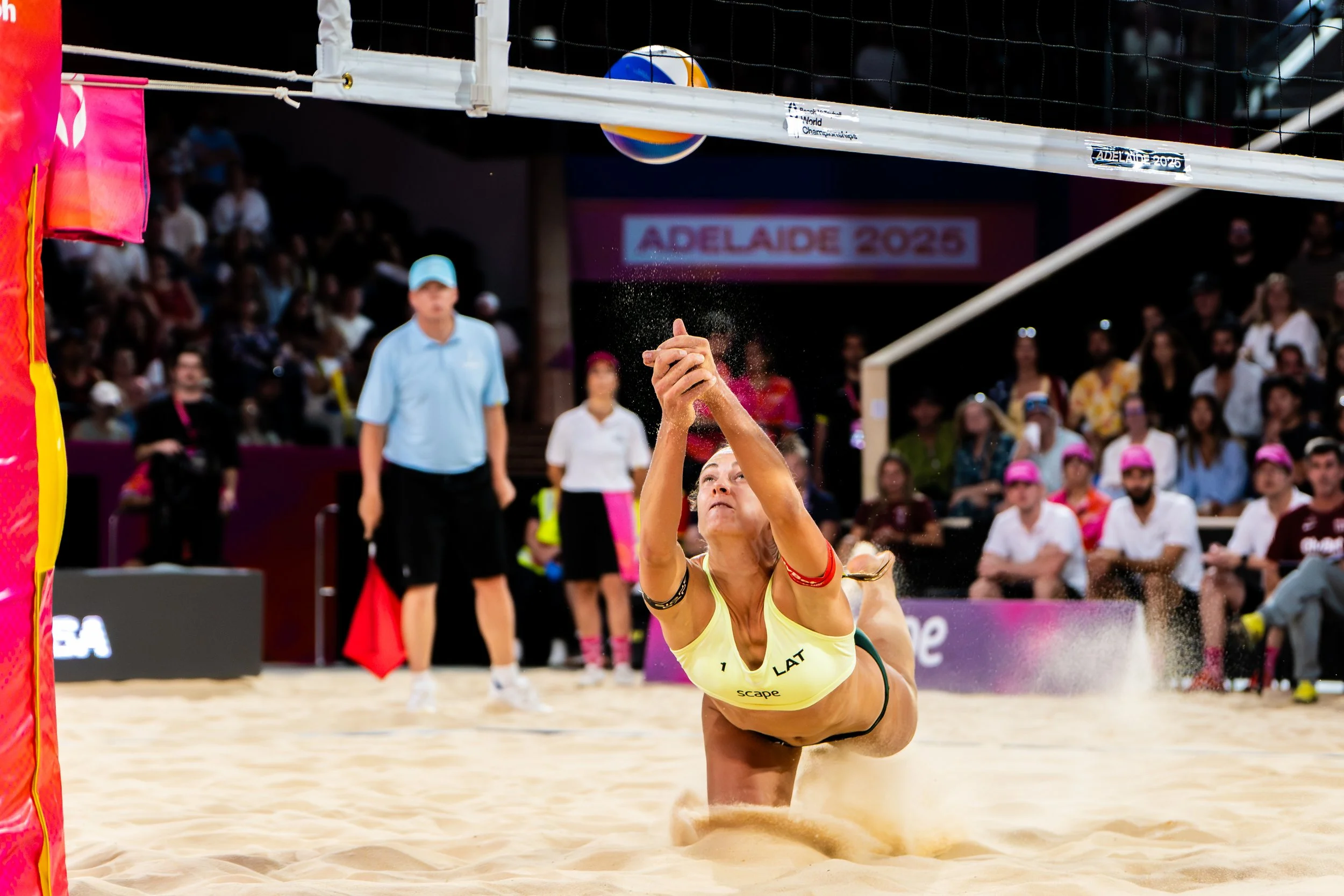 A female beach volleyball player in a yellow sports bra and black shorts diving into the sand attempting to save the ball during a match at the FIVB 2025 World Beach Volleyball Championships, with spectators and officials watching in the background.