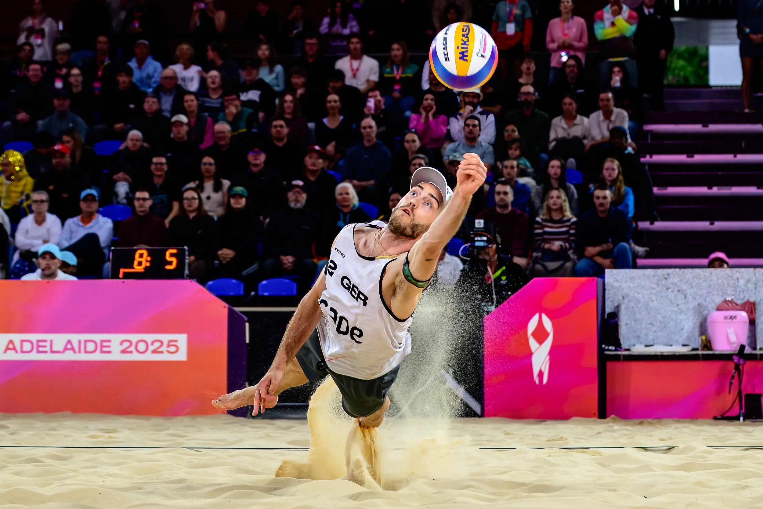 A male beach volleyball player diving to hit the ball during a match at the 2025 FIVB World Beach Volleyball event, with a crowd of spectators in the background.
