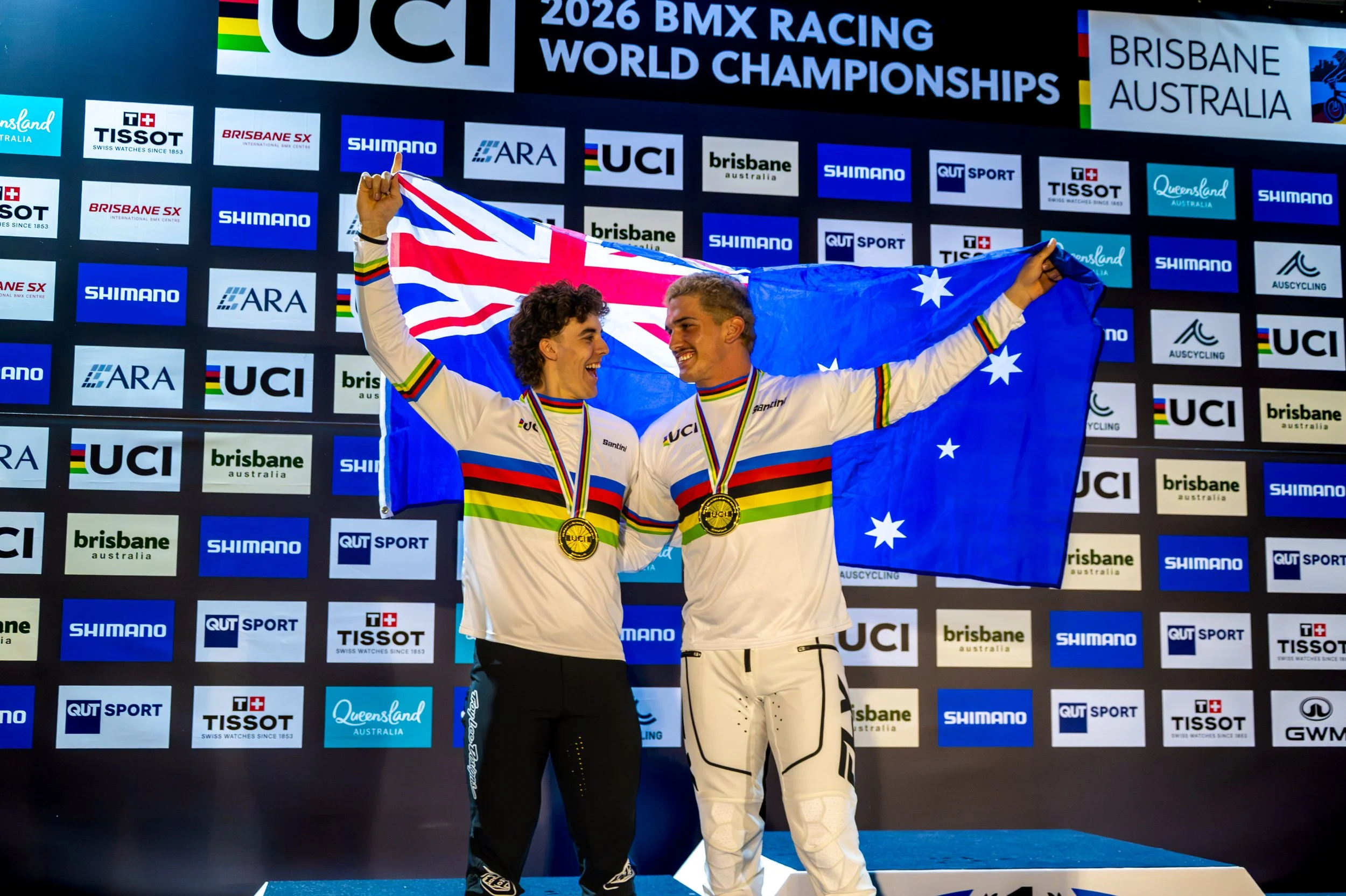 Two athletes in racing suits celebrating on the winners' podium, holding an Australian flag, with medals around their necks in front of a backdrop displaying various sponsors and event information at the 2026 BMX Racing World Championships in Brisban