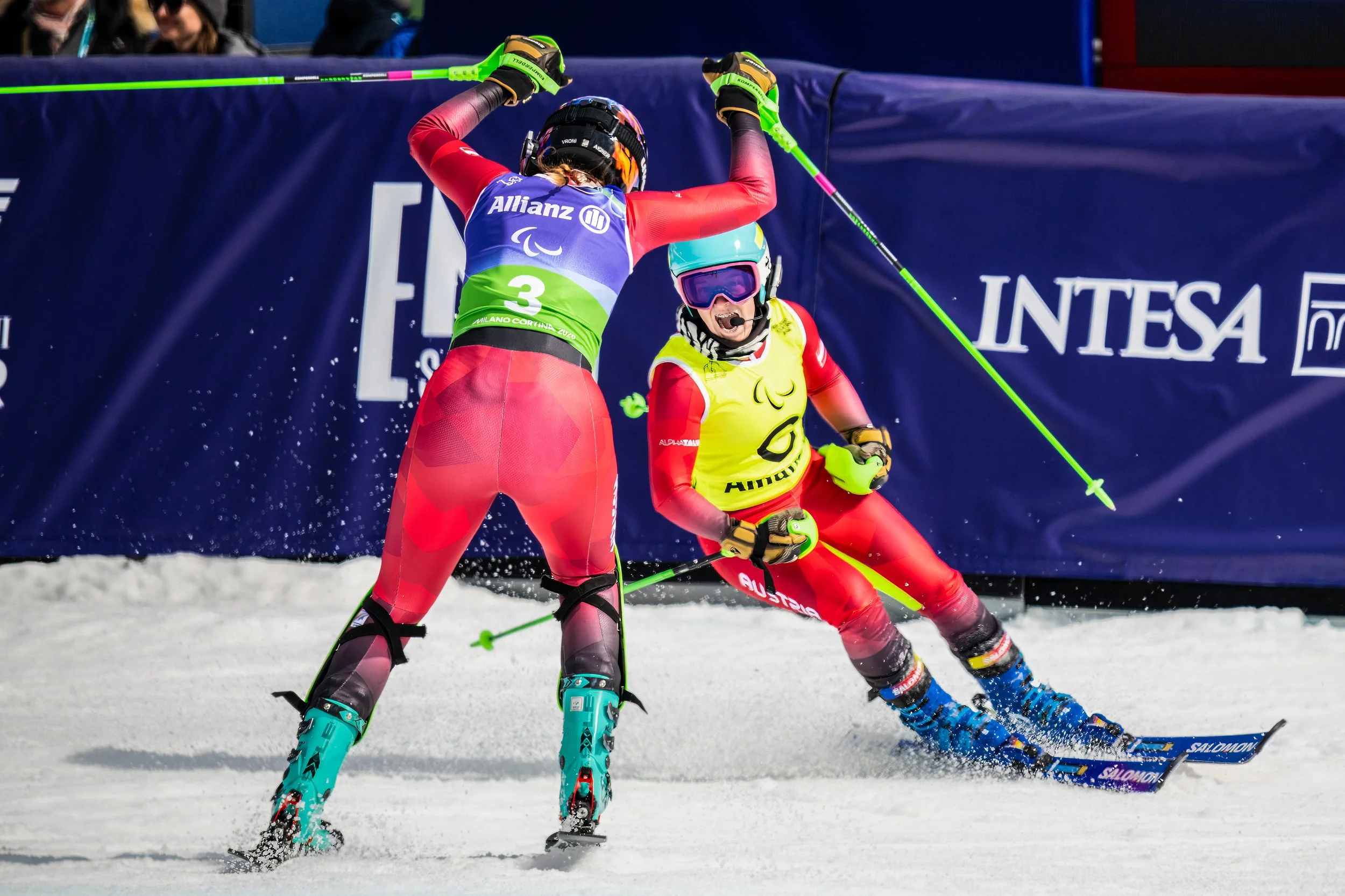 Two skiers celebrating at the 2026 MilanoCortina Winter Paralympics in snowy terrain, wearing helmets, goggles, and colorful race suits during a ski event, with a blue banner in the background.