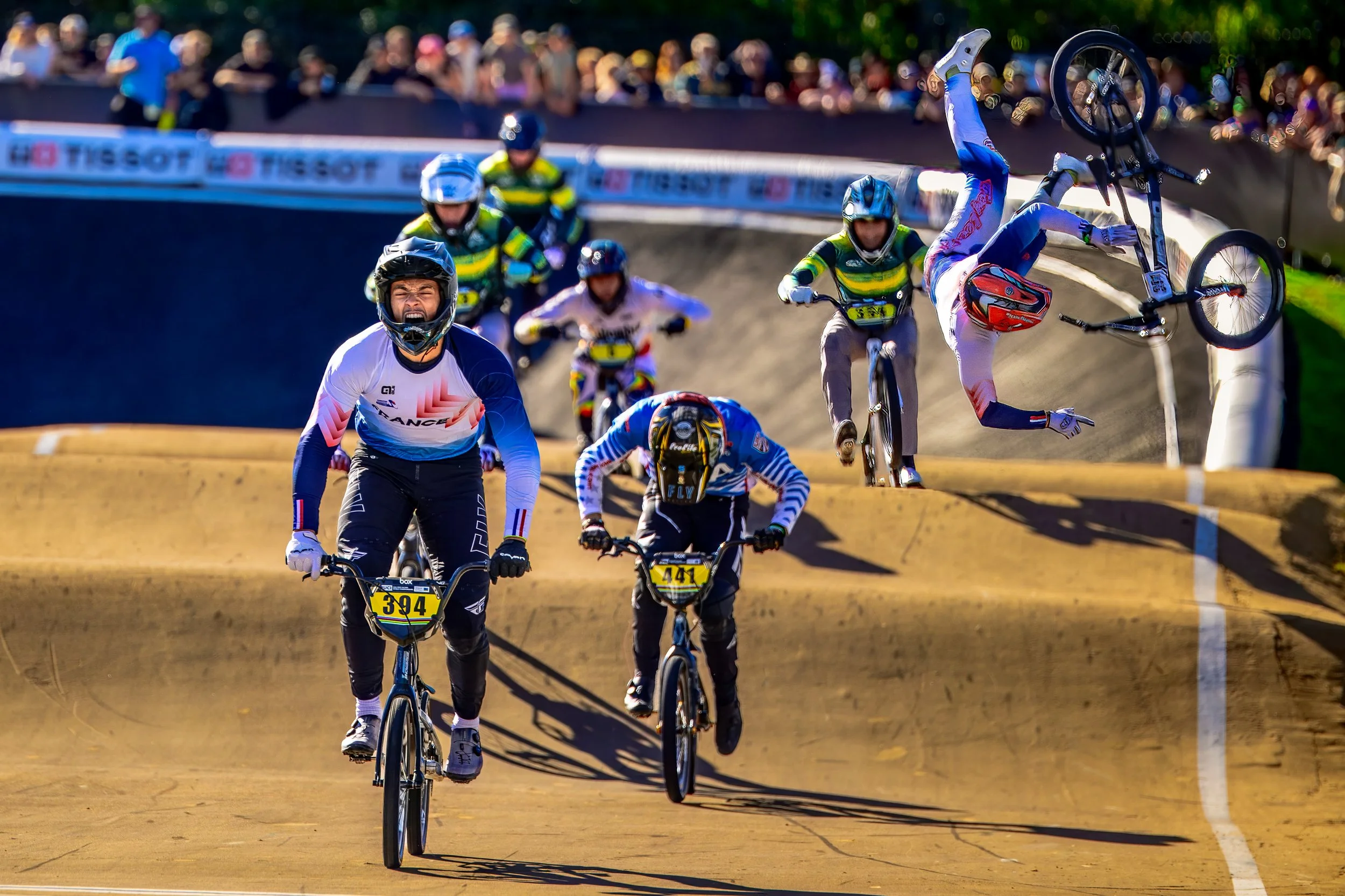 BMX riders competing in a race at the 2026 UCI BMX World Championships, with one rider performing a flip and others cycling behind, and spectators watching from the sidelines.