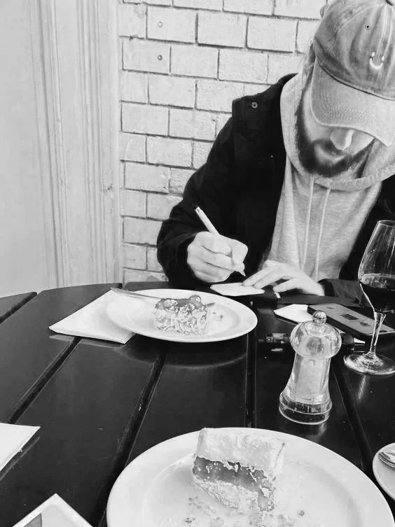 Ein Mann mit Mütze zeichnet in einem Restaurant, im Vordergrund ein Stück Kuchen auf einem Teller, auf dem Tisch Salz- und Pfefferstreuer, ein Glas Wein und Papieren.