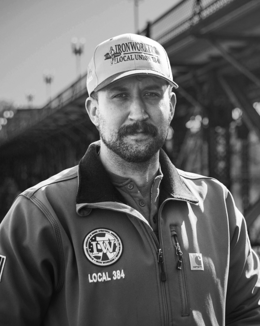 A man with a mustache and beard wearing a baseball cap, jacket, and work uniform stands outdoors near a bridge, with a focused expression.