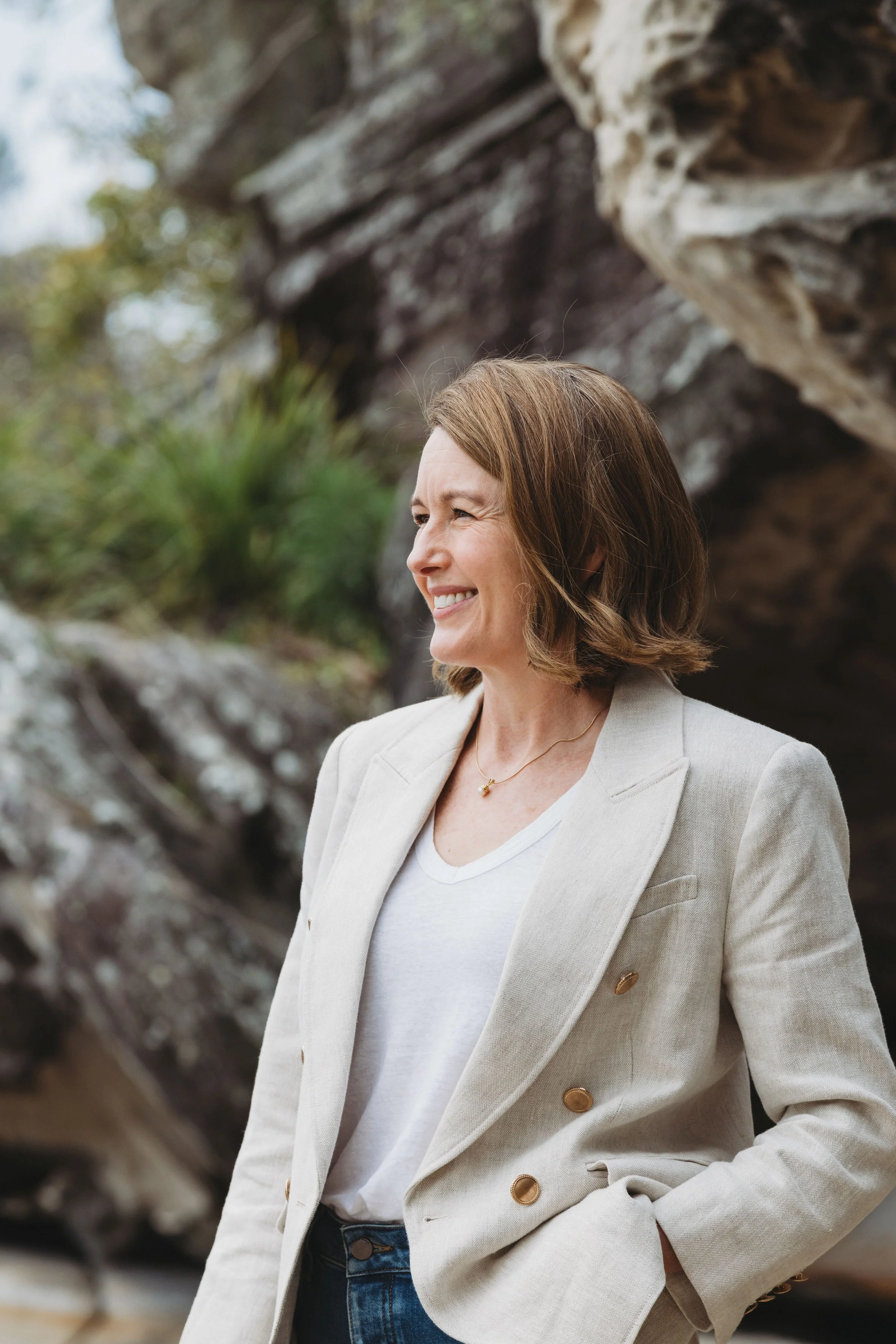 A woman with shoulder-length brown hair smiling, wearing a beige blazer over a white T-shirt, with a background of rocky landscape and some blurred greenery.