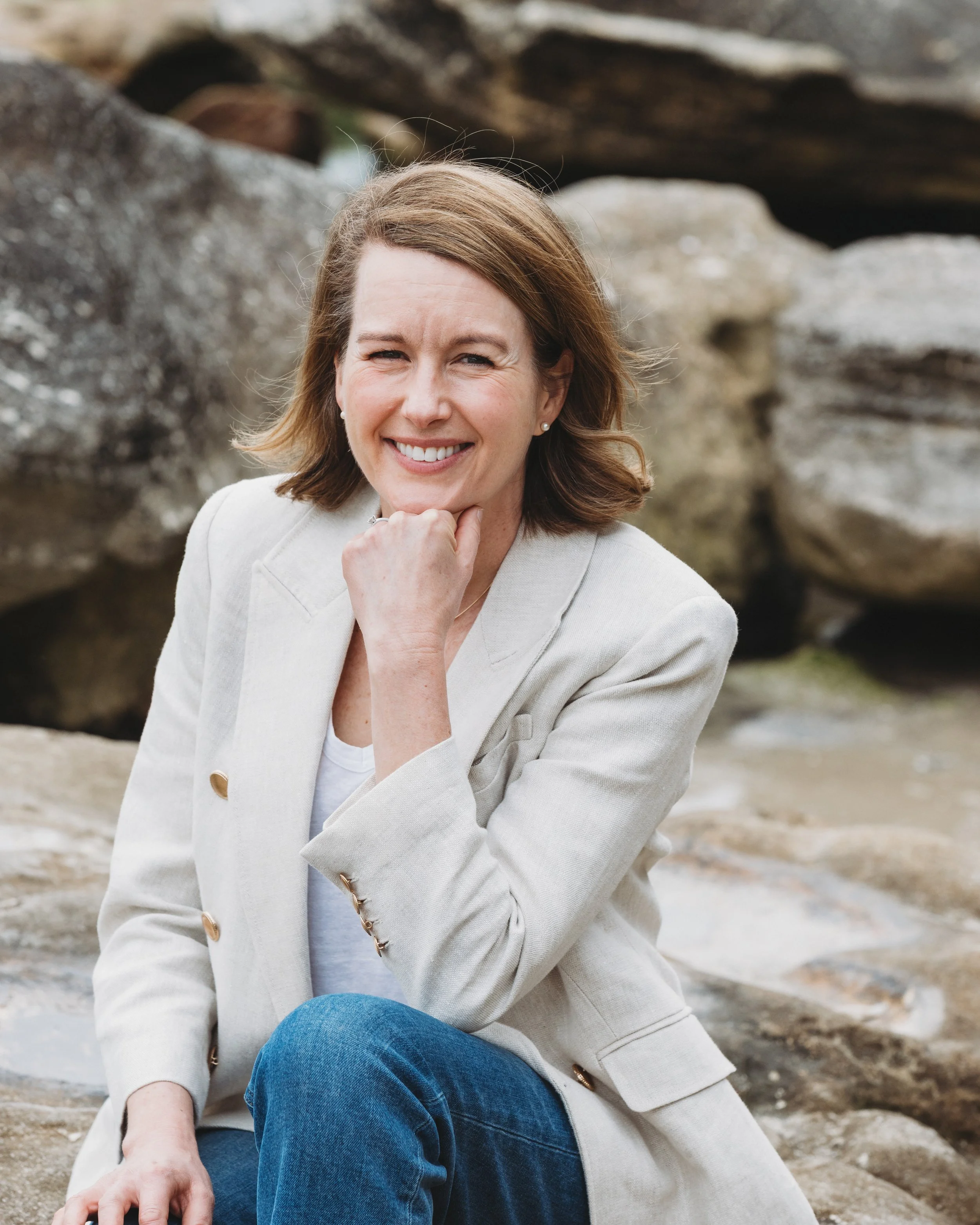 A woman with light brown hair, wearing a light-colored blazer and blue jeans, sitting outdoors on a rocky surface and smiling at the camera.