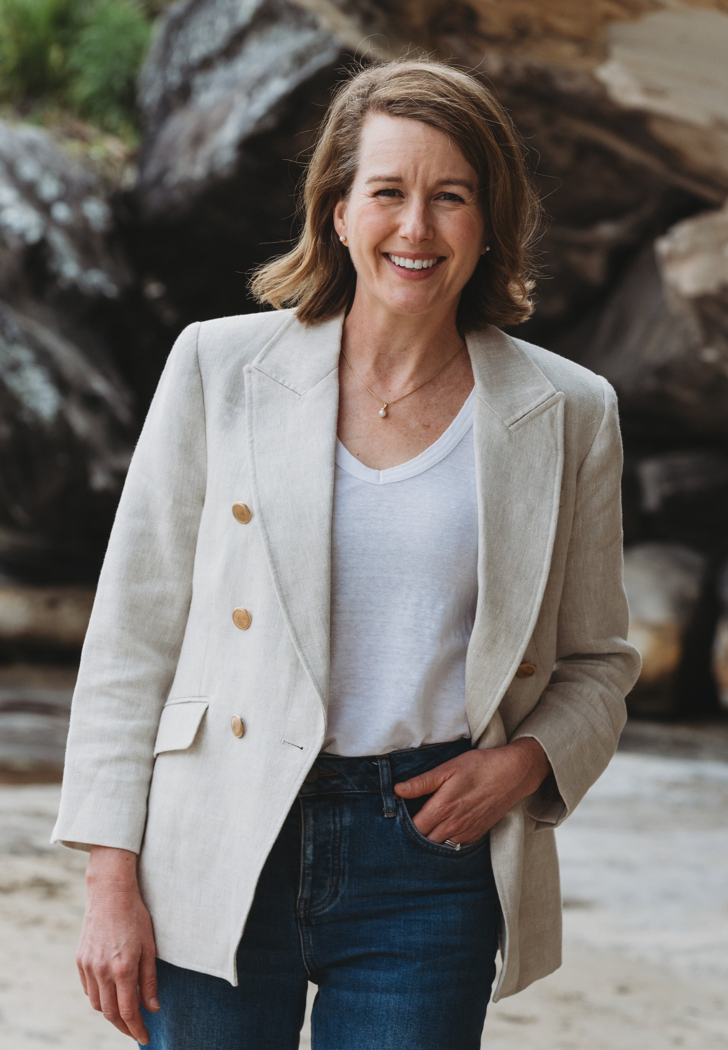 A woman with shoulder-length brown hair smiling outdoors, wearing a light-colored blazer over a white T-shirt and dark jeans, with a rocky background.