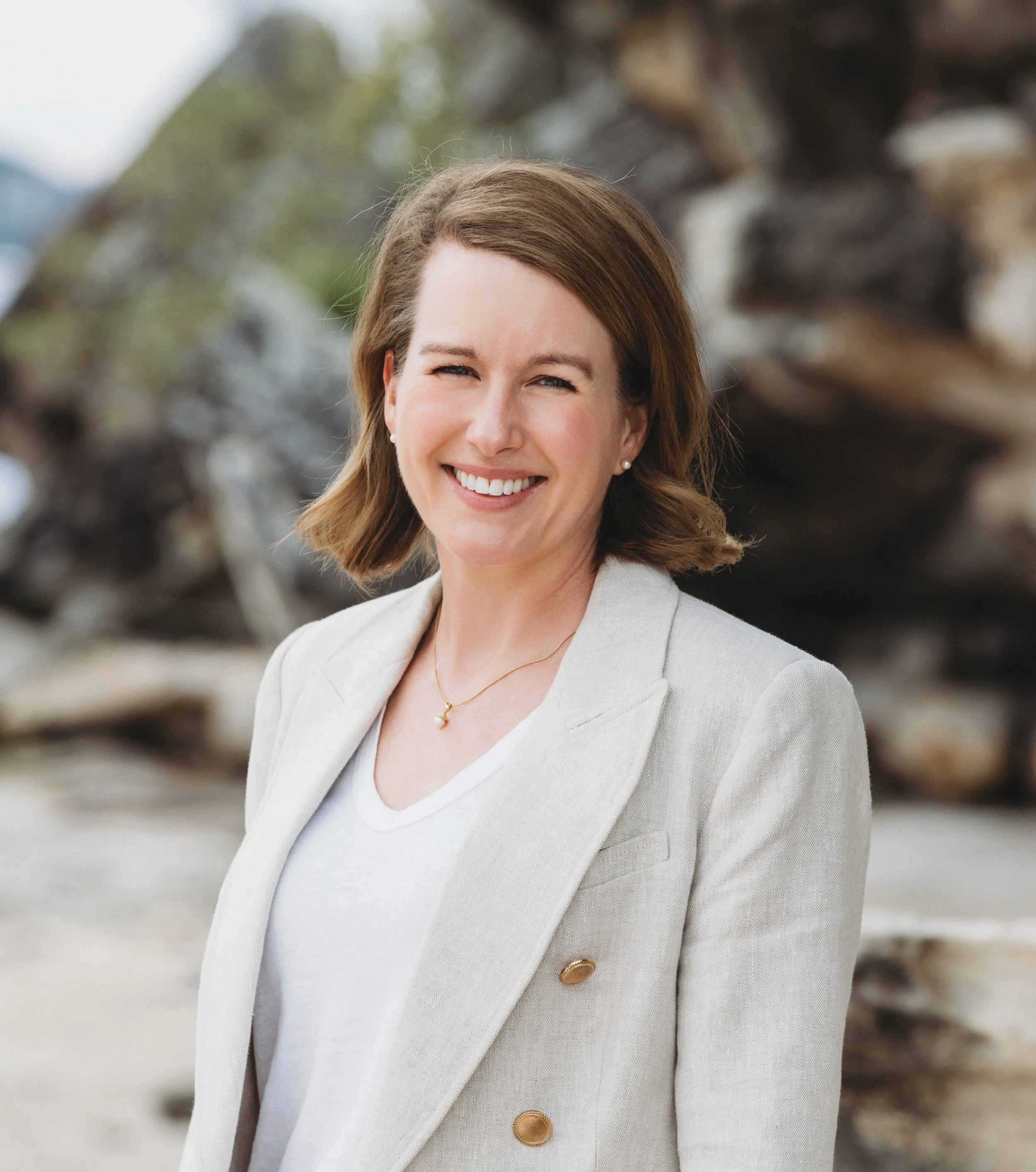 A woman with shoulder-length brown hair wearing a light-colored blazer and white shirt, smiling outdoors with a blurred background of rocks and trees.