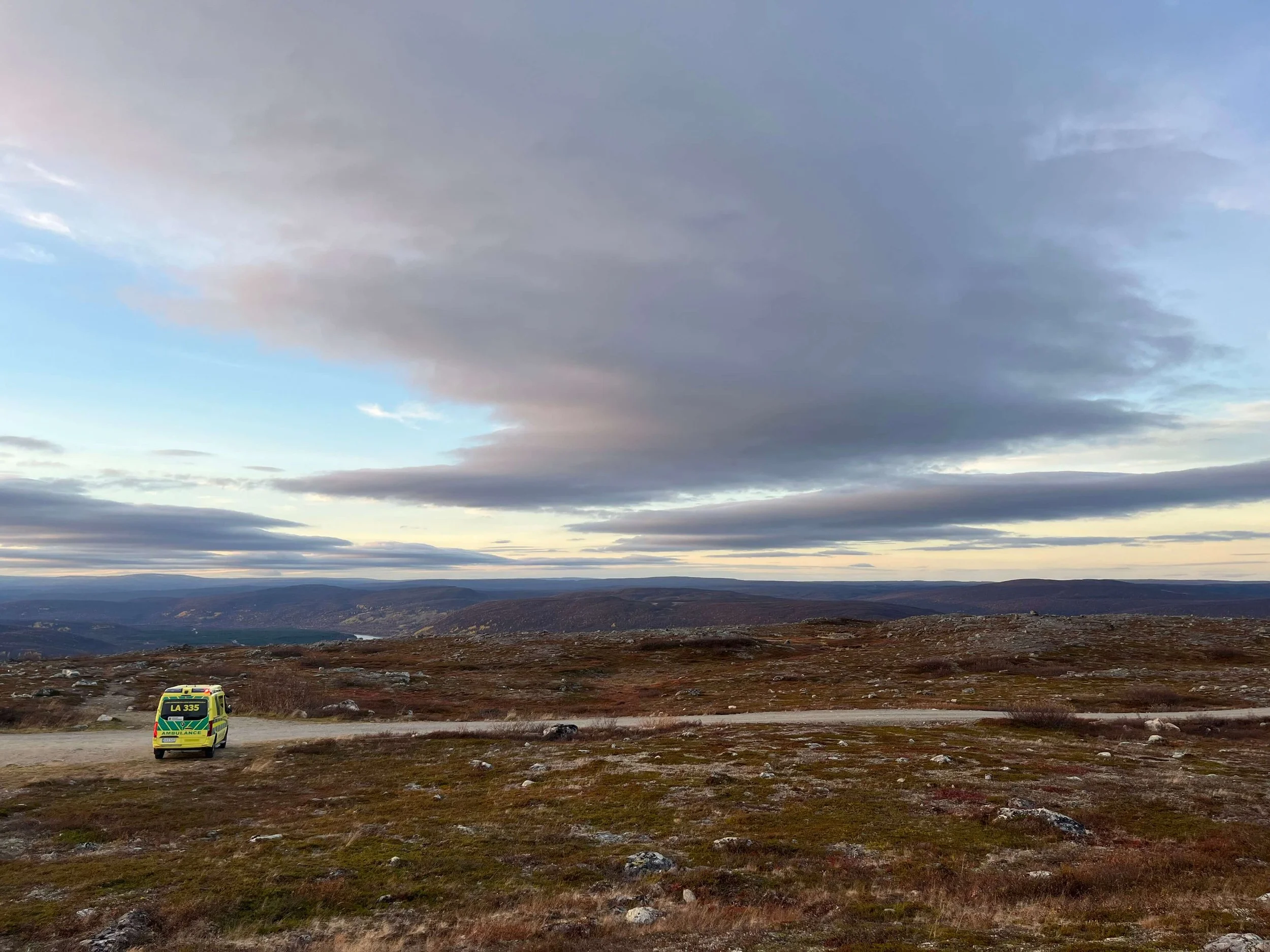 A yellow ambulance parked in the countryside, with a flat landscape and a cloudy sky in the background.