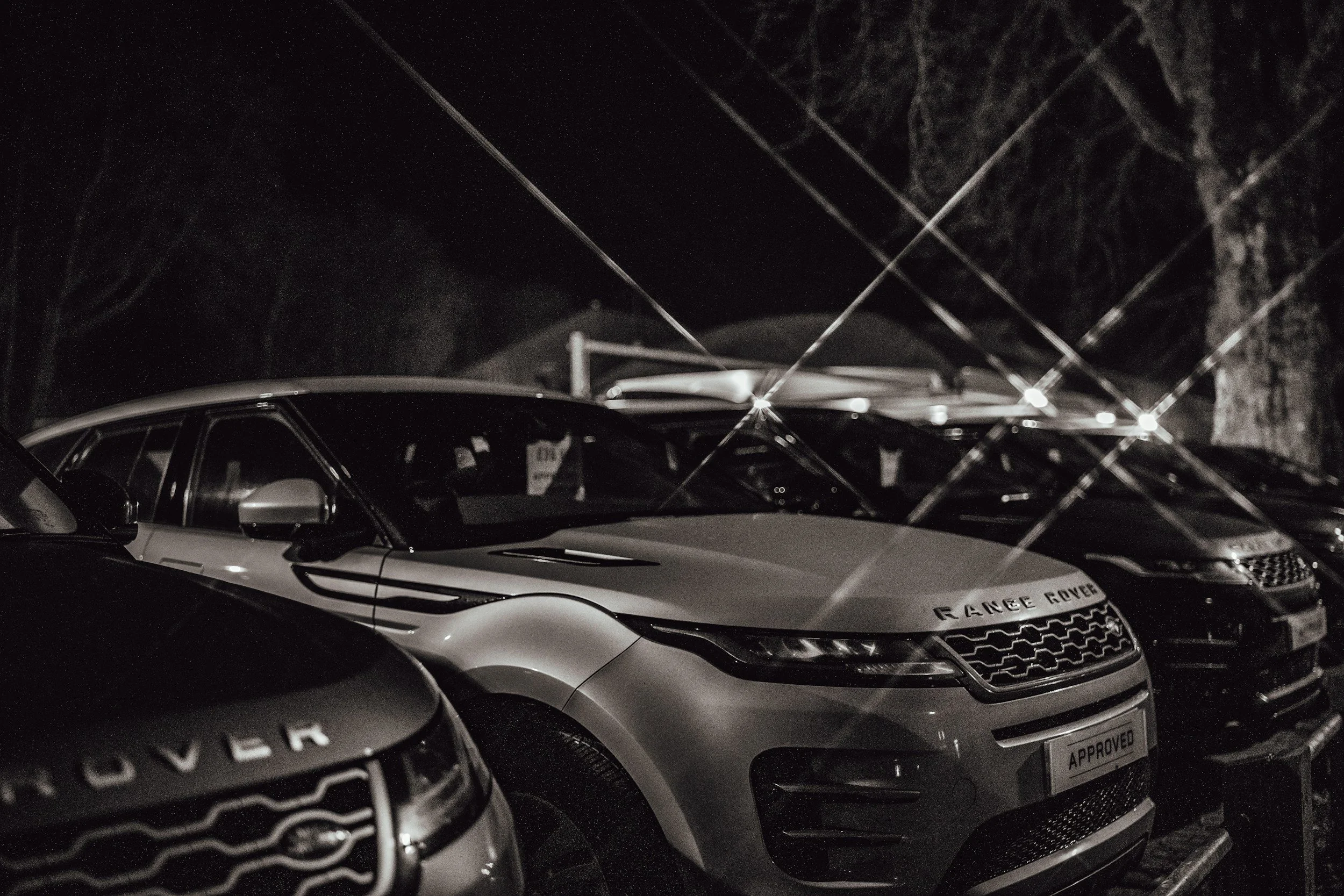 A row of parked cars at night, illuminated by bright lights reflecting off the vehicles and creating starburst effects.