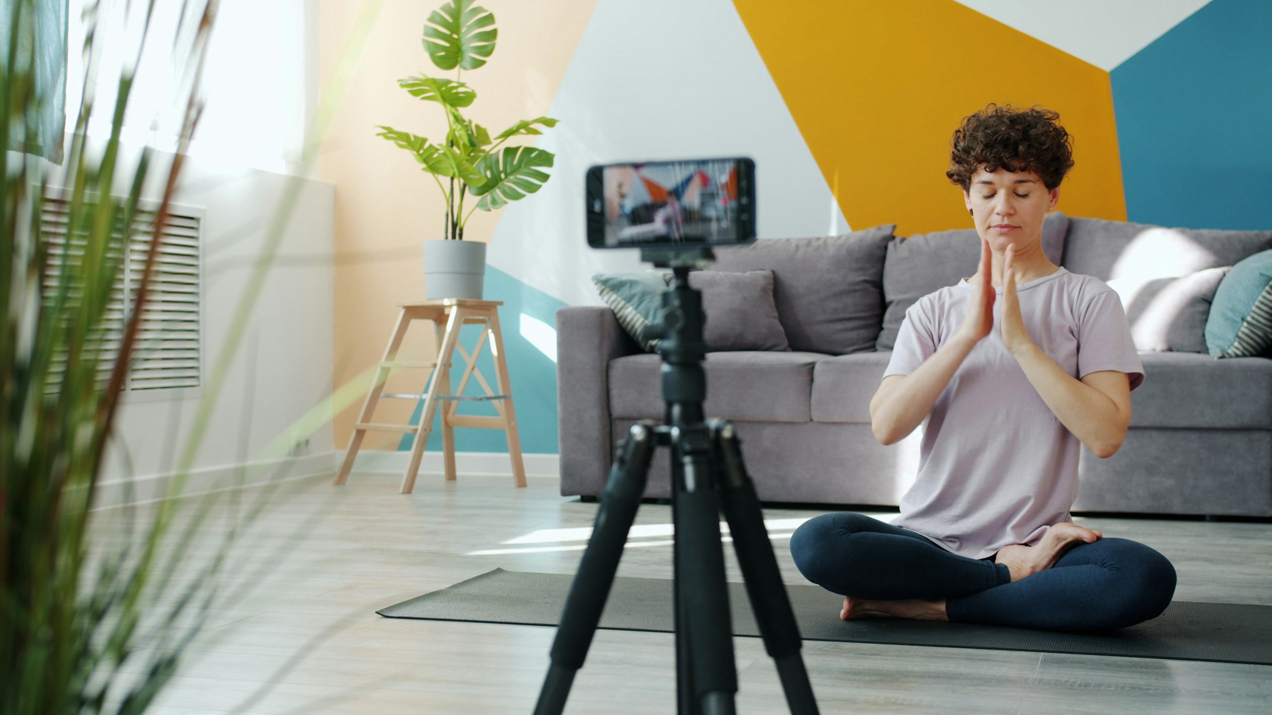 A person with curly hair sitting cross-legged on a yoga mat in a living room, meditating with hands in a prayer position, in front of a camera on a tripod capturing the practice.