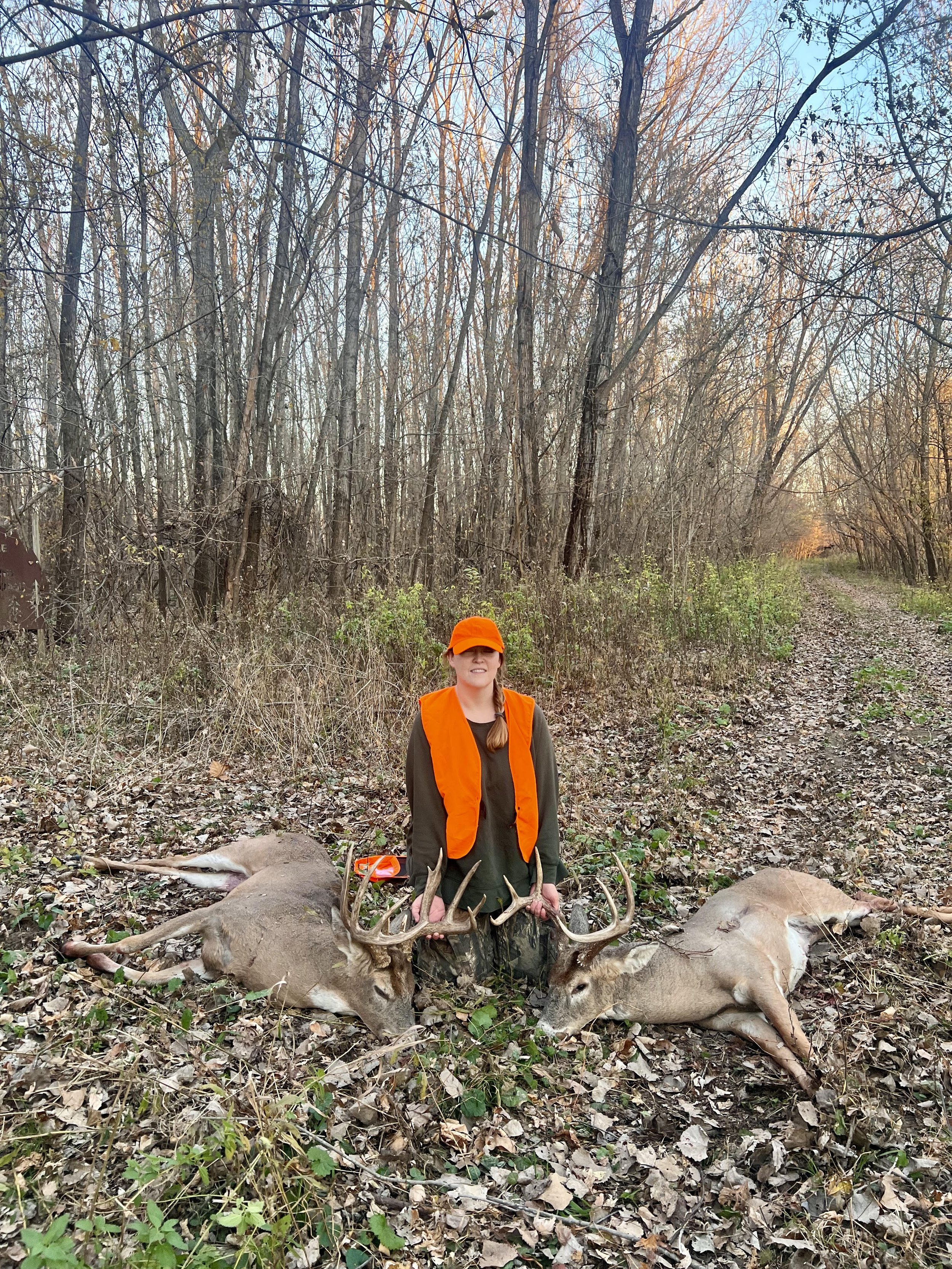 Hunter kneeling between two harvested whitetail bucks on a wooded trail while wearing hunter orange at Tegridy Outdoors in Missouri.