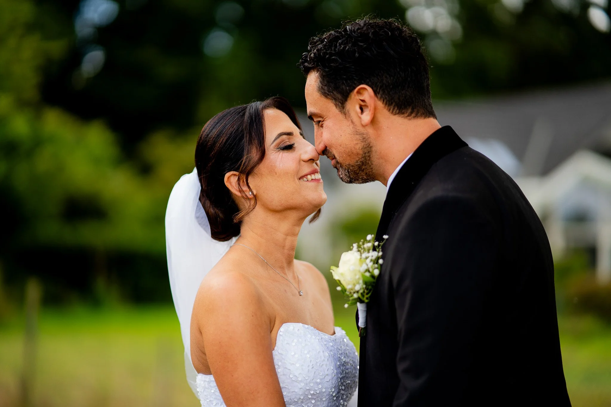 Ein frisch vermähltes Brautpaar steht sich bei ihrer Hochzeit gegenüber, mit der Stirn aneinander, mit einem Lächeln. Sie trägt ein weißes, trägerloses Brautkleid und eine Schleife im Haar. Er trägt einen schwarzen Anzug. Im Hintergrund sind grüne Bä