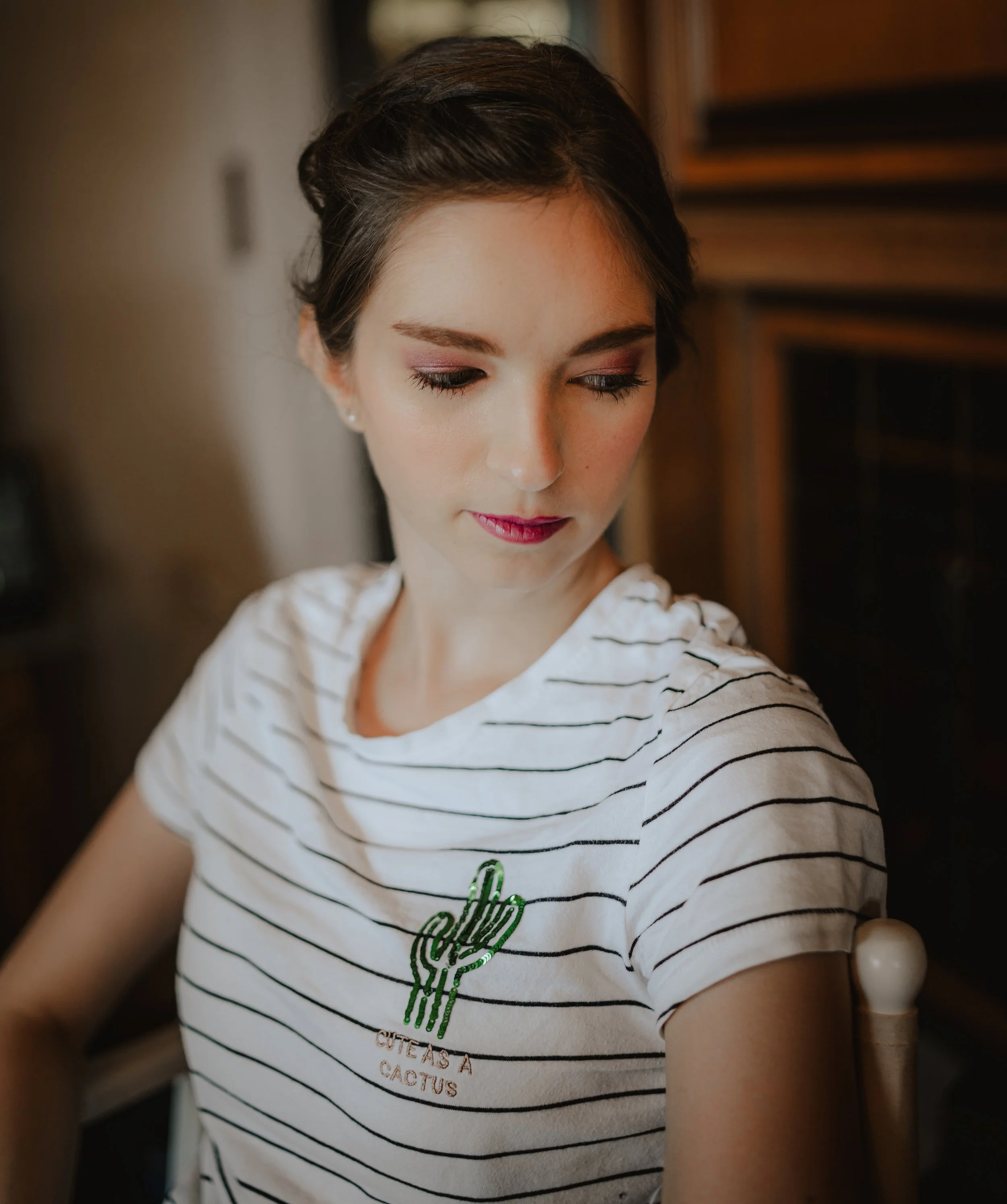 Junge Frau mit dunklem Haarknoten, Makeup, in weißen T-Shirt mit schwarzen Streifen und Kaktus-Design, sitzt in einem Raum mit Holzregal im Hintergrund.