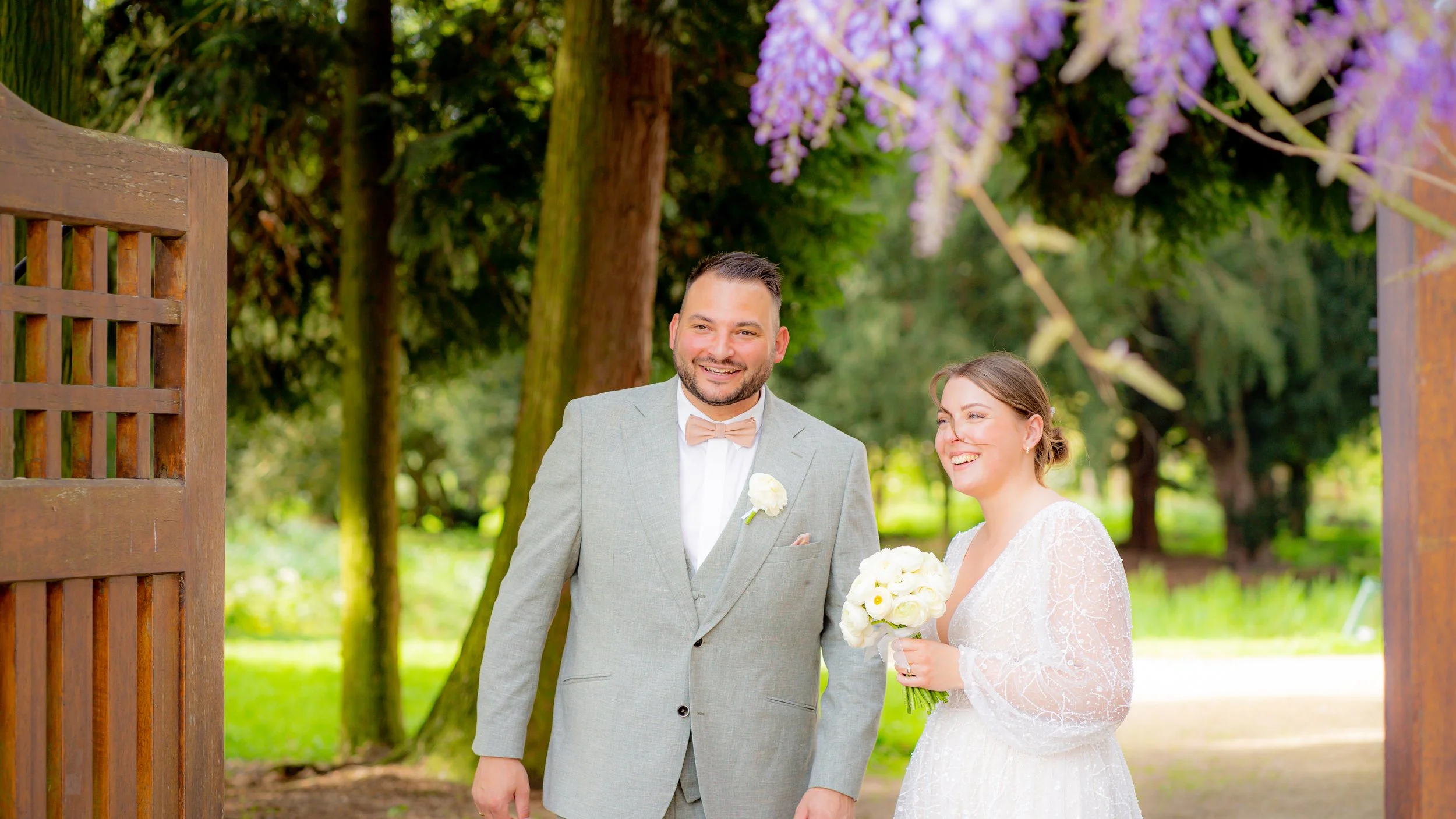 Ein lachendes Brautpaar bei ihrer Hochzeit im Freien, umgeben von grünen Bäumen und blühenden lila Fliederbüsche, mit der Braut in einem weißen Kleid und dem Bräutigam in einem hellgrauen Anzug.