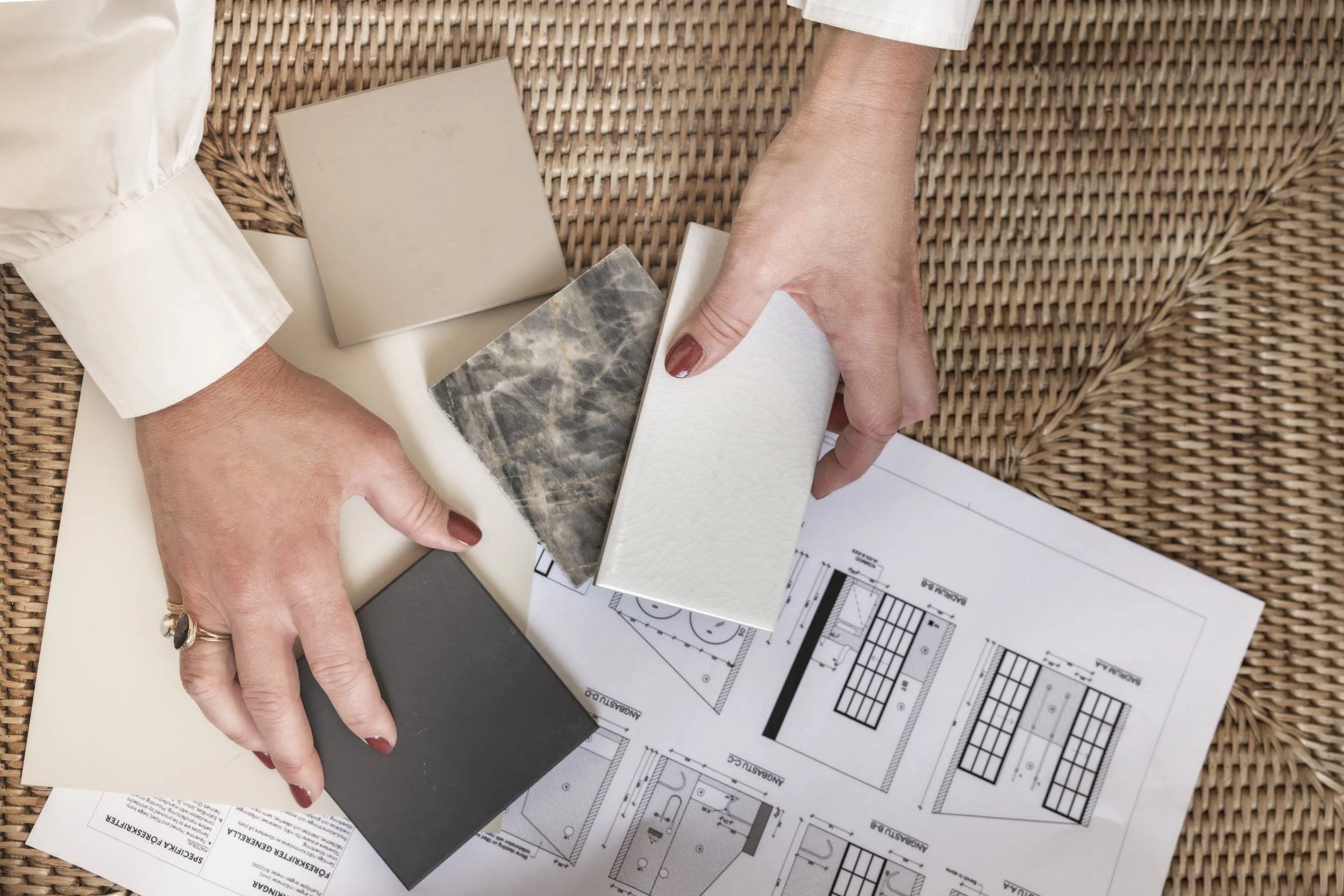 Hands arranging sample tiles on architectural blueprints on a woven surface.