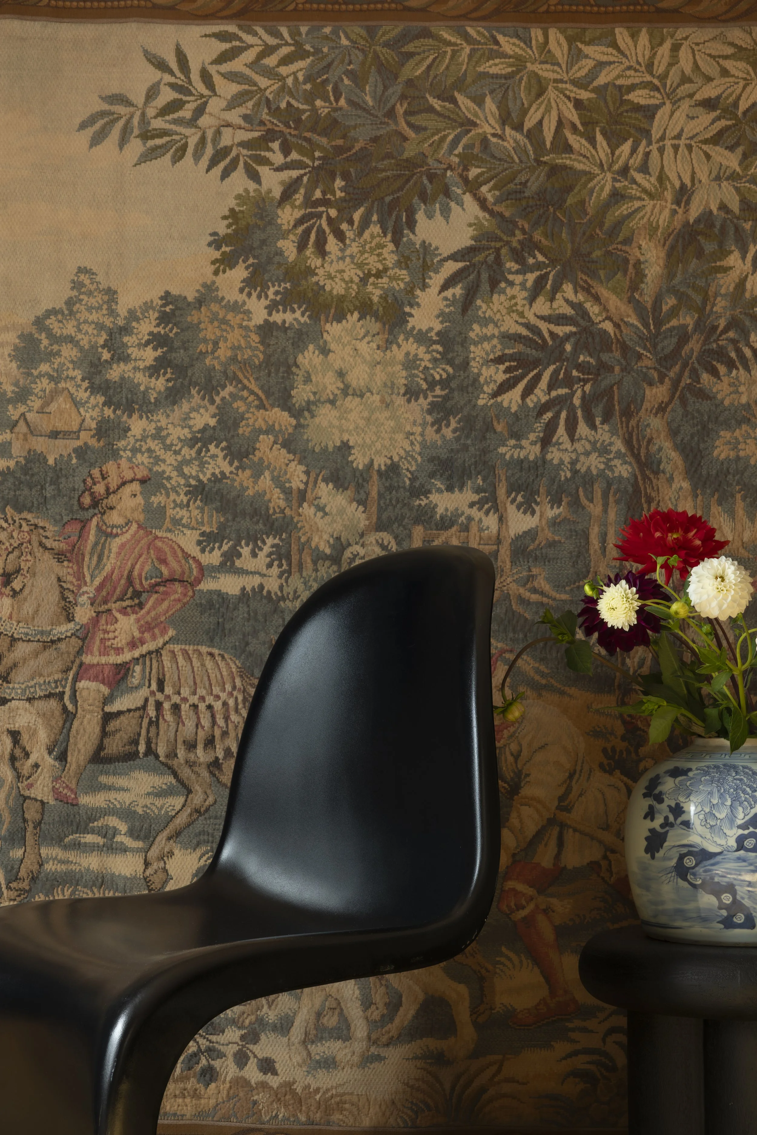A black modern chair in front of a vintage tapestry depicting a pastoral scene with trees and a person riding a horse. To the right, there is a blue and white ceramic vase with red, white, and dark red flowers on a small round table.