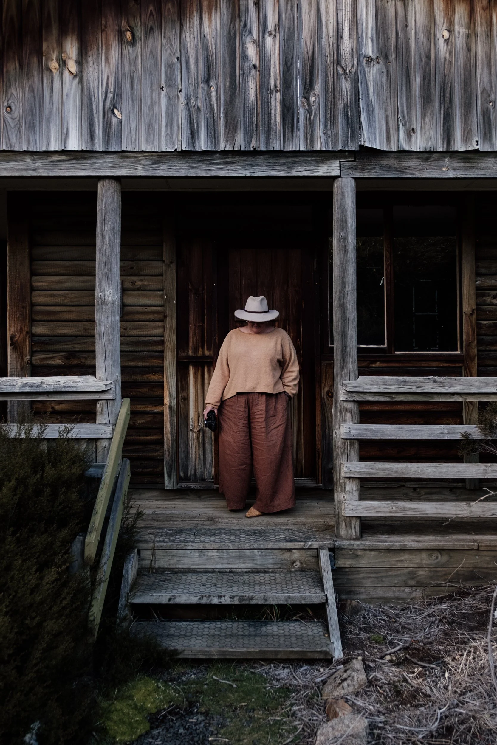 Woman standing on a wooden porch in front of a rustic house holding a camera