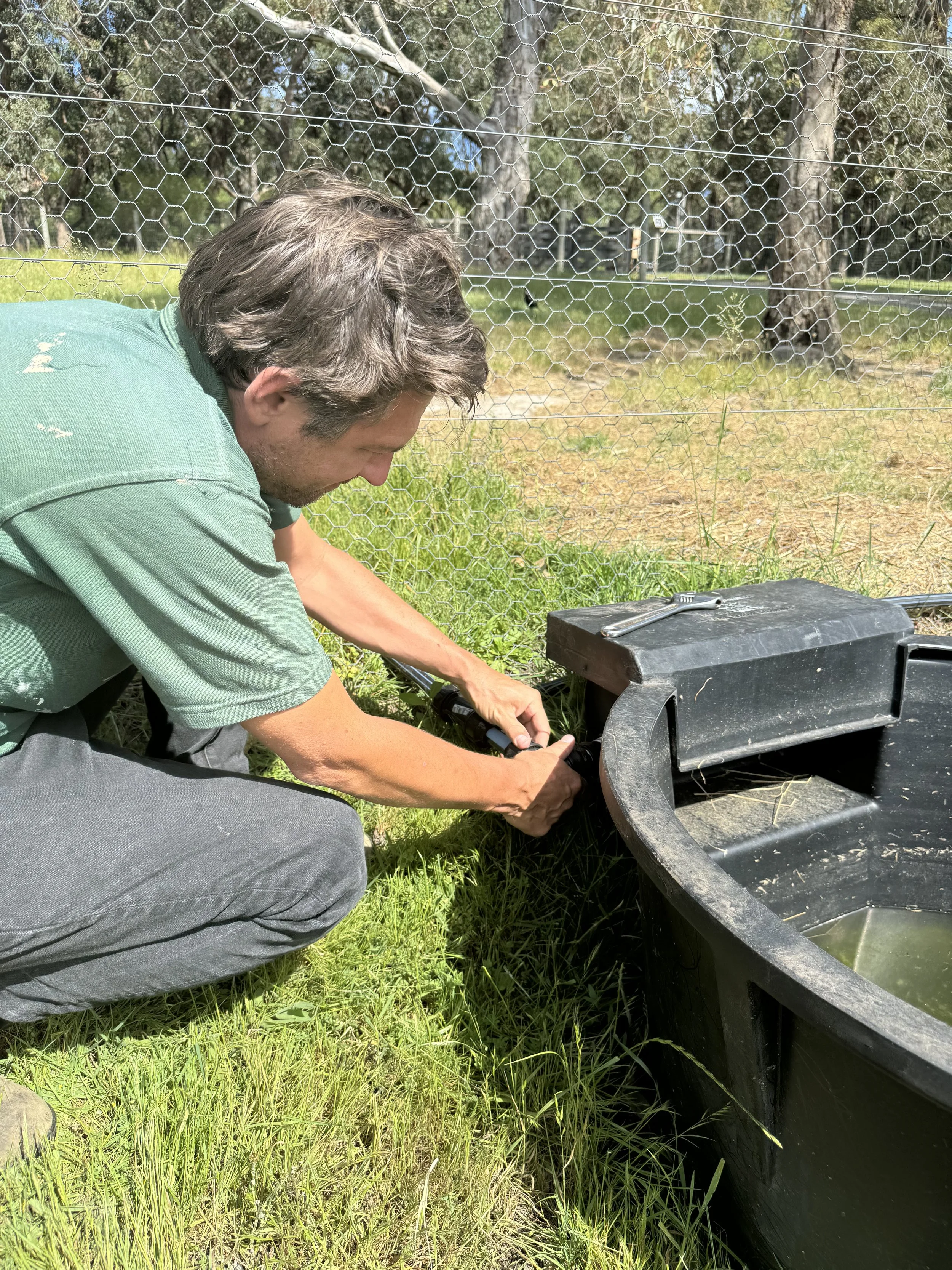 A man crouching next to a black water tank, using a wrench on a large bolt, outside in a grassy area enclosed by wire fencing.