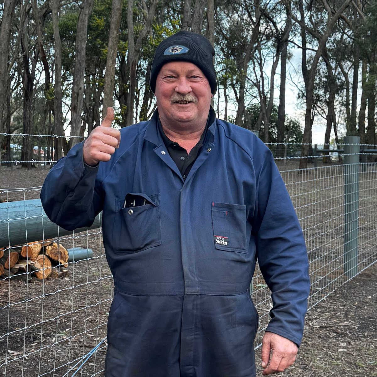 A middle-aged man with a mustache giving a thumbs-up and smiling outdoors, wearing a black beanie and a blue work jumpsuit, standing near a wire fence with trees in the background.