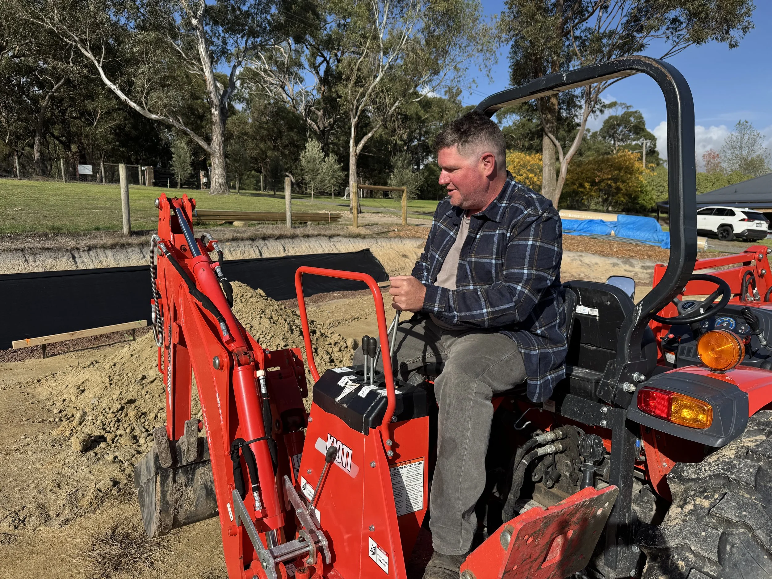 A man operating a red compact excavator at a construction site on a sunny day with trees and parked cars in the background.