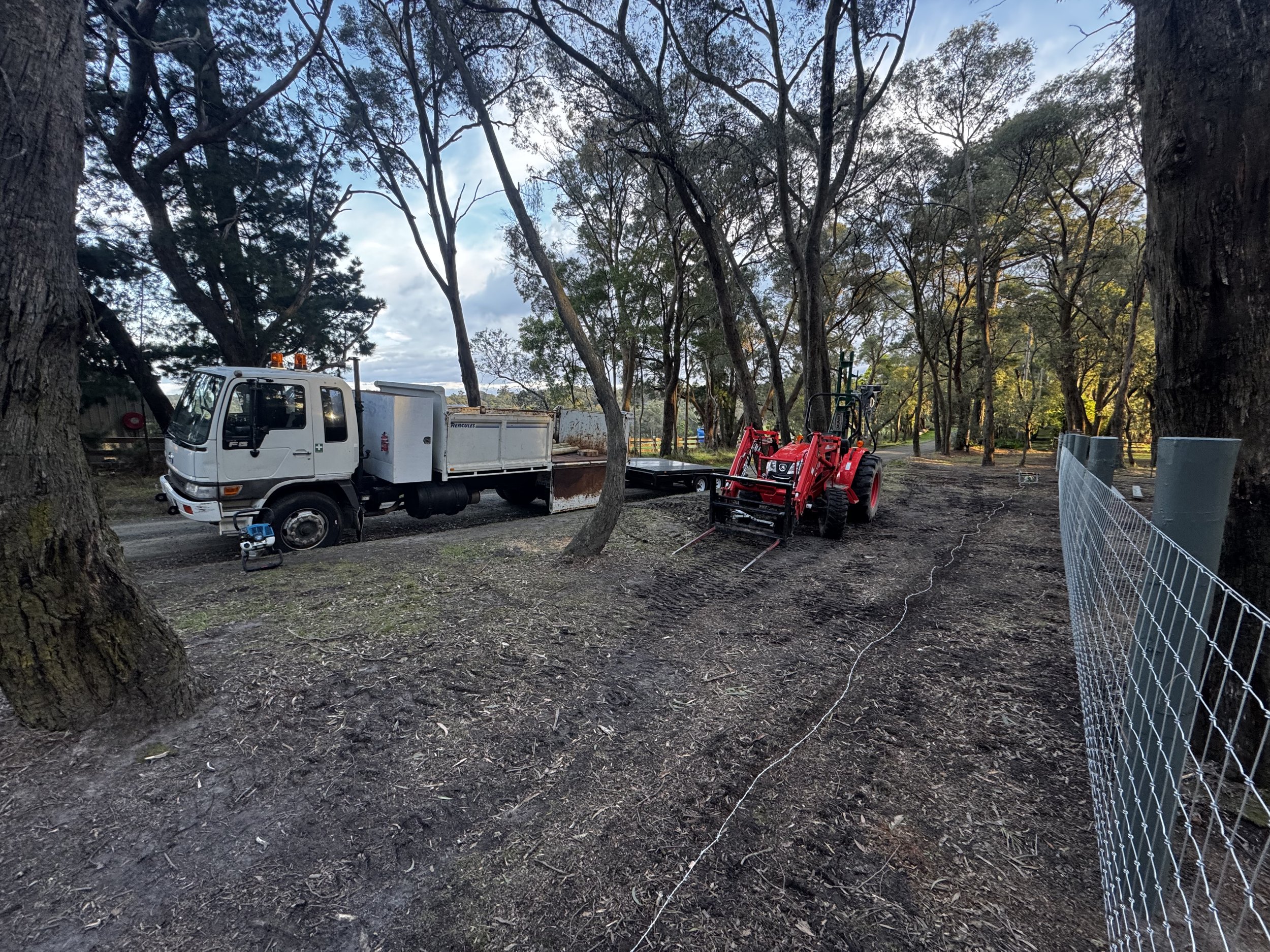 A worksite with a white utility truck and a red tractor surrounded by trees and fencing, with dirt ground.