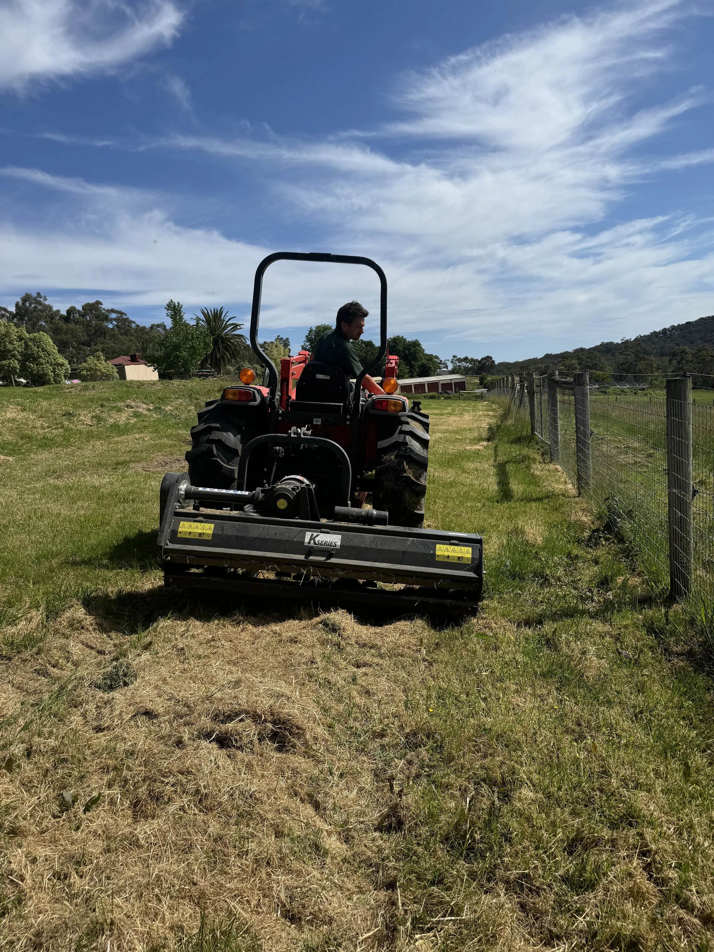 A man operating a small red tractor with a mower attachment, cutting grass alongside a wire fence in a rural area with trees, hills, and a partly cloudy sky in the background.