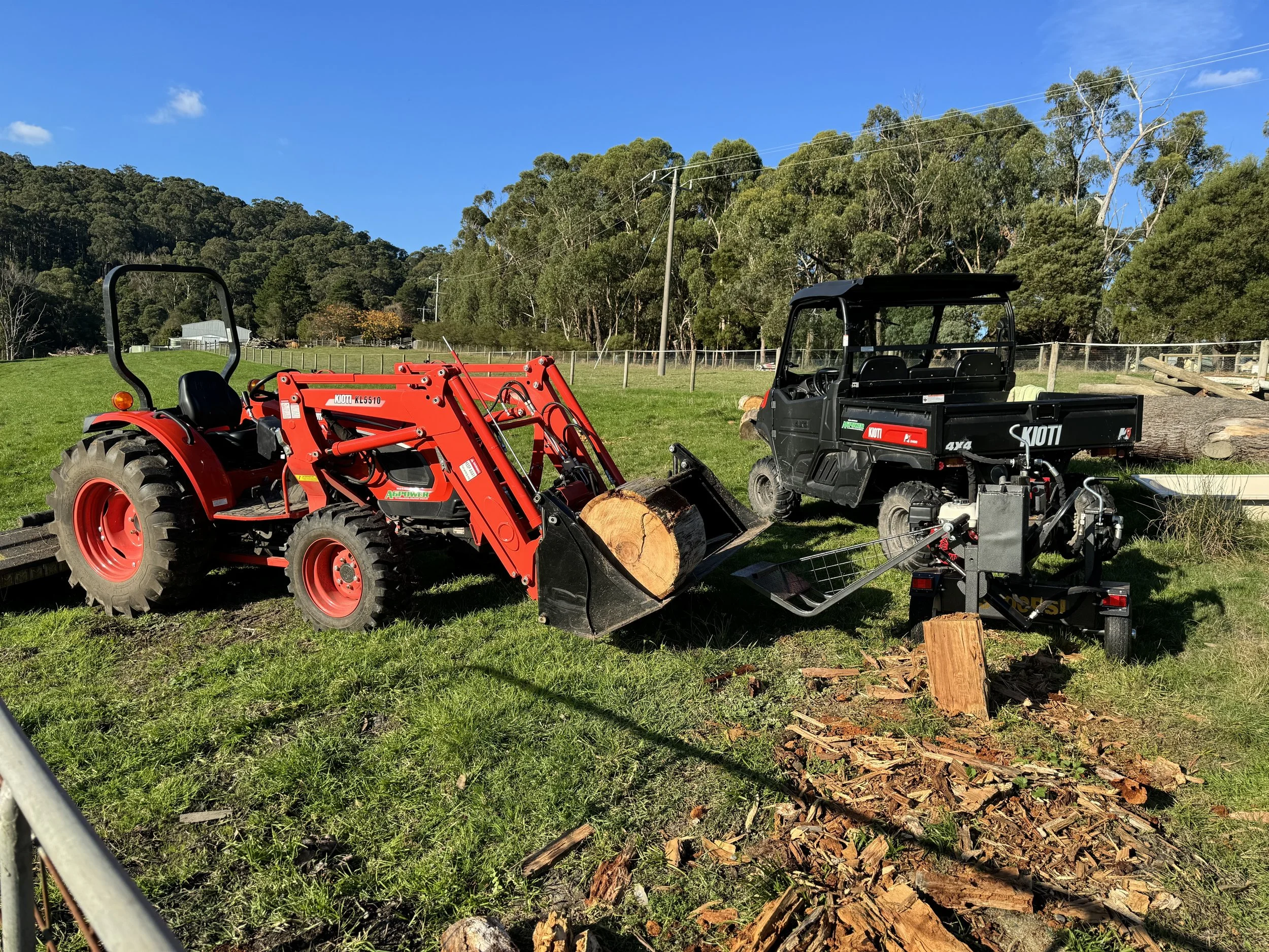 A tractor and a log splitter on a grassy field, with logs and wood debris nearby, trees and a fence in the background, and a clear blue sky above.