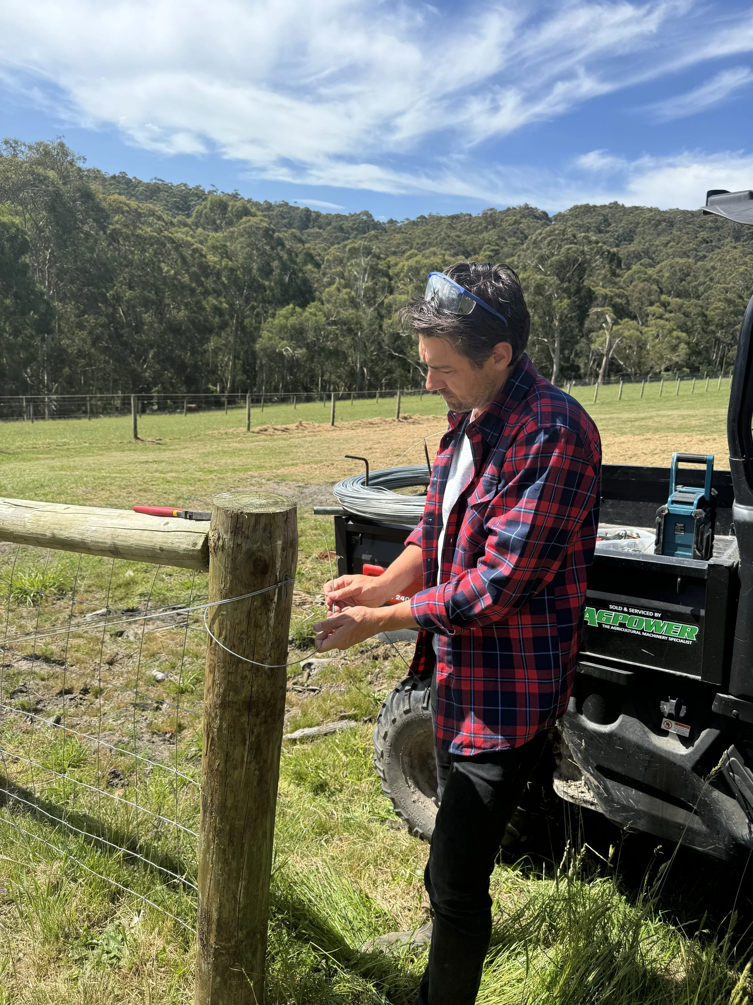 A man in a red plaid shirt and sunglasses working on a wire fence in a grassy field with trees and hills in the background under a blue sky with clouds.