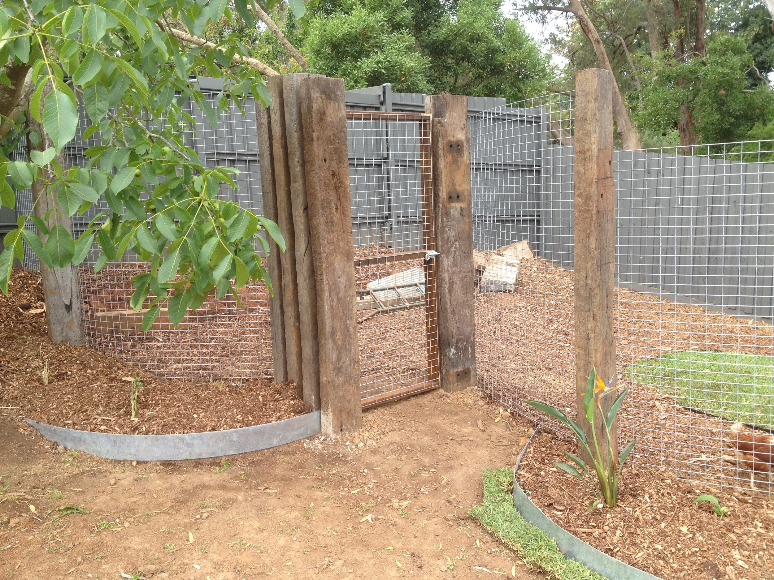 An outdoor animal enclosure with wire fencing and wooden posts, containing dirt, plants, and a small animal area.